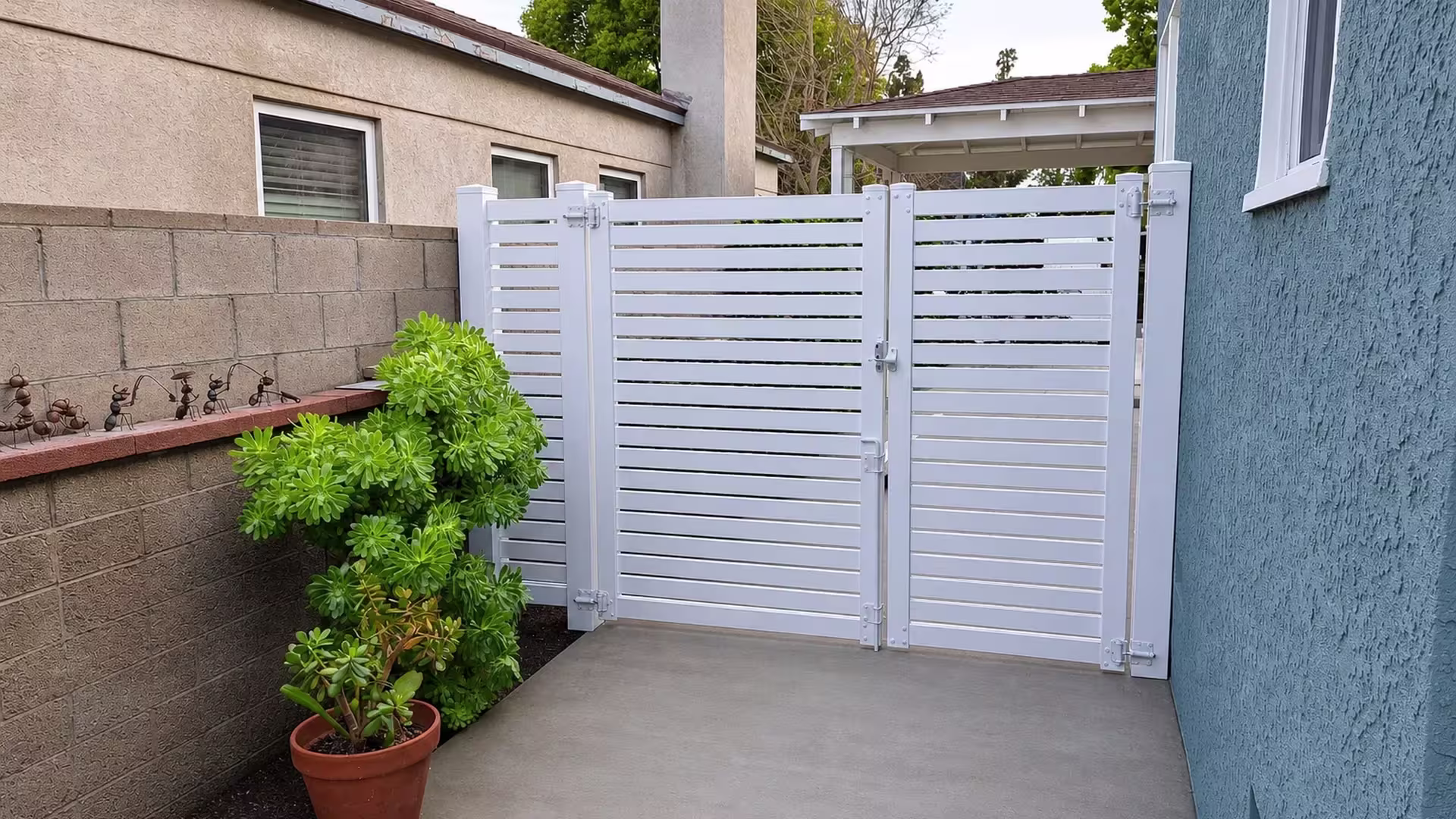 Succulent shrubs and a potted plant sit beside this White vinyl slatted driveway gate between two buildings