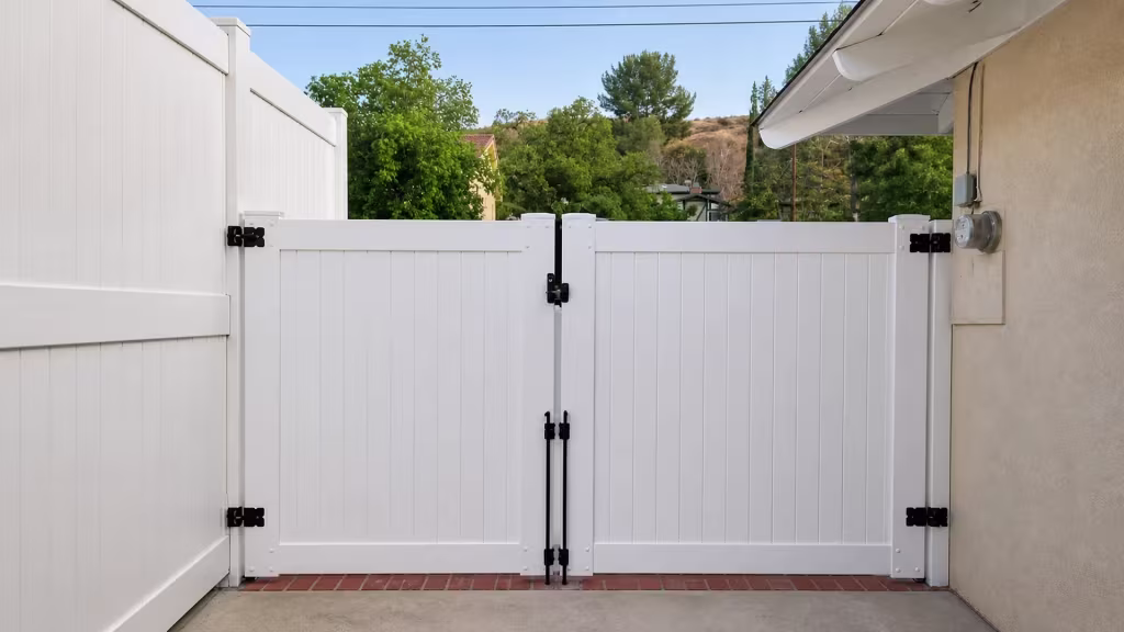 Vertical tongue-and-groove slats face this White vinyl driveway gate as its two leaves swing open over a brick threshold