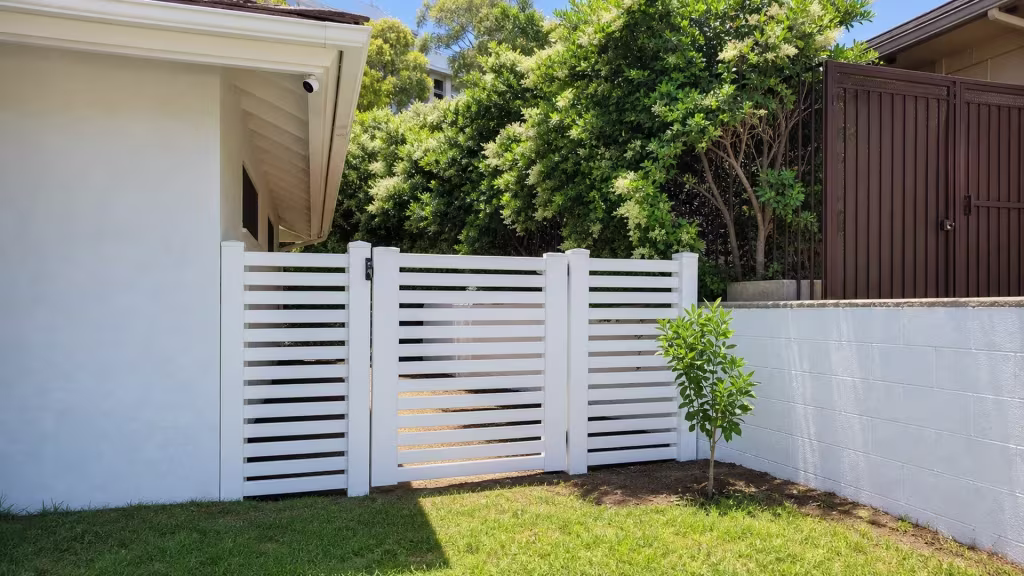 This White vinyl driveway gate spans from the house wall to a White masonry wall in three graduated sections