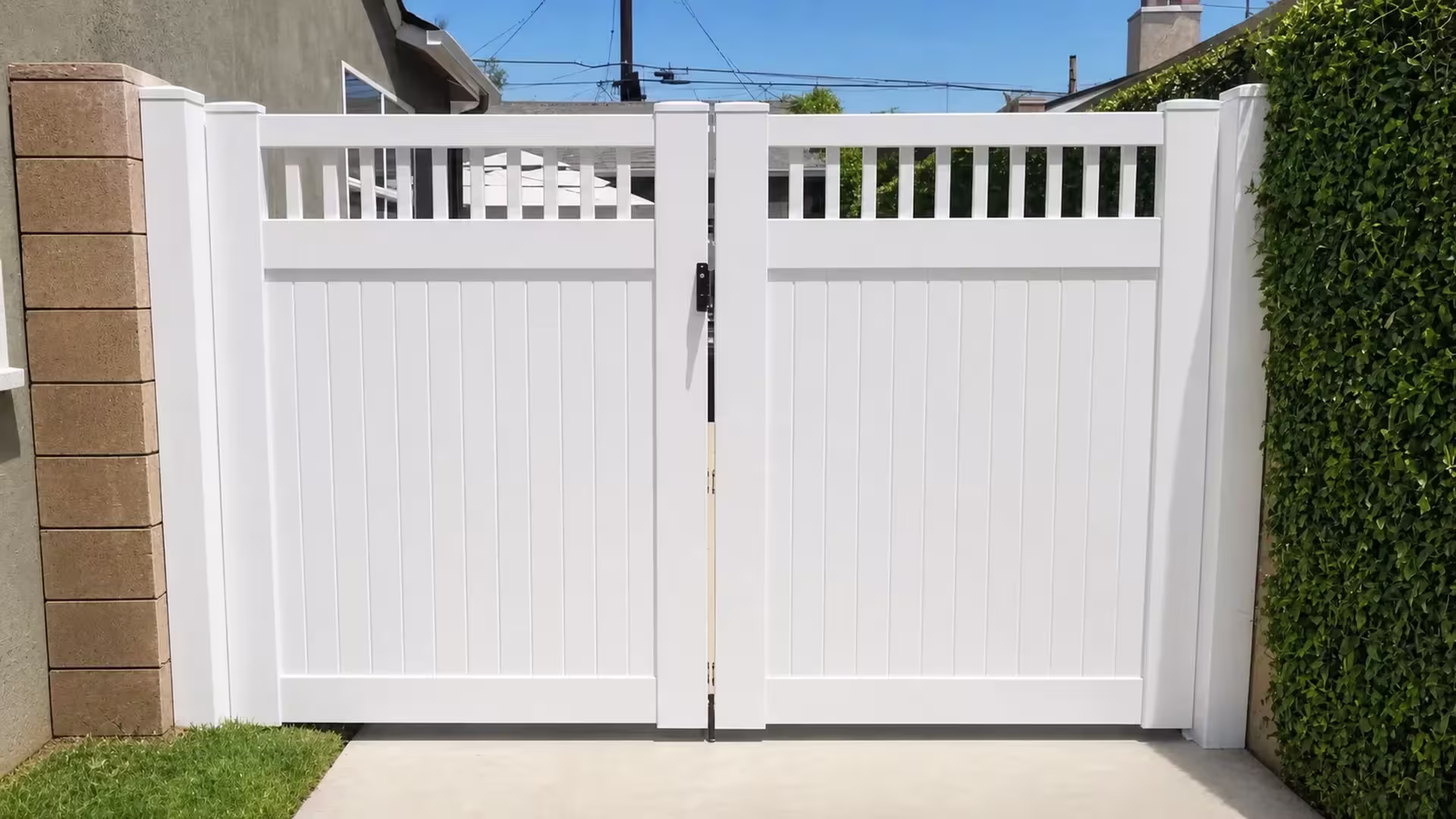 White vinyl driveway gate closing between a masonry house wall and an ivy-covered wall on the other side