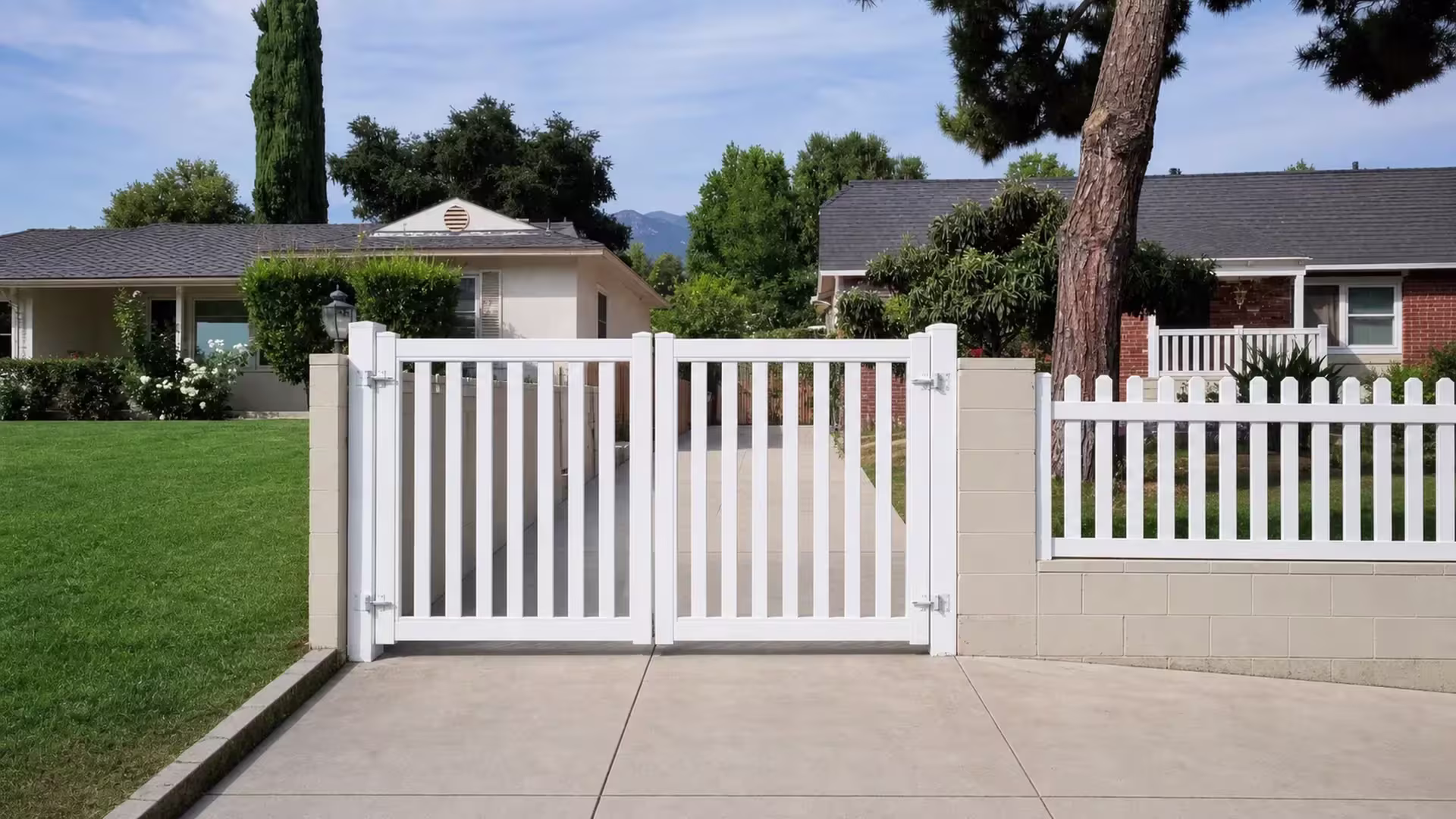 White vinyl double-swing driveway gate standing along the drive beneath a mature pine canopy