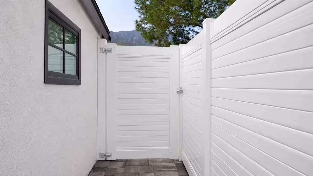 White vinyl horizontal-privacy gate crosses a paved side yard, with a mature pine tree and a distant mountain beyond