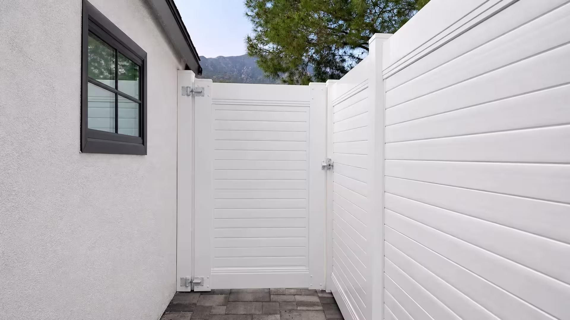 White vinyl horizontal-privacy gate crosses a paved side yard, with a mature pine tree and a distant mountain beyond