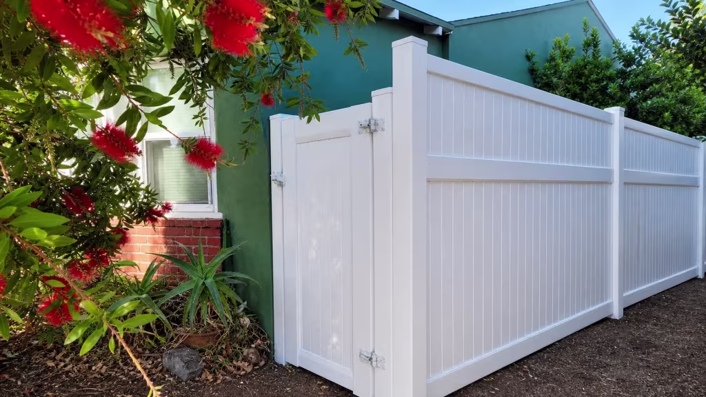 White vinyl privacy gate stands beside a green house wall, its brick planter edge holding aloe and a bottlebrush tree