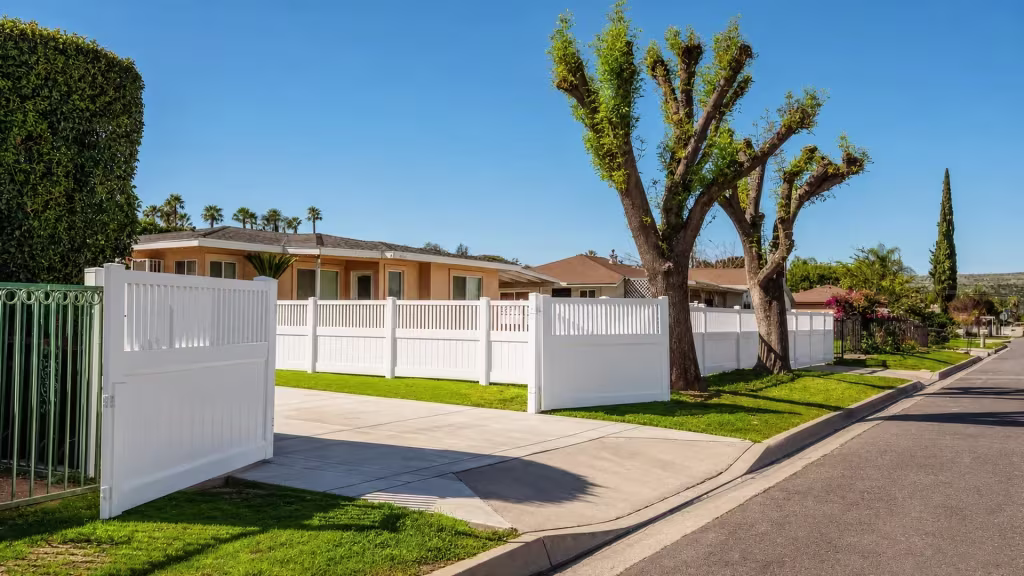 White vinyl driveway gate swung open toward the street beside a green metal fence at the property line