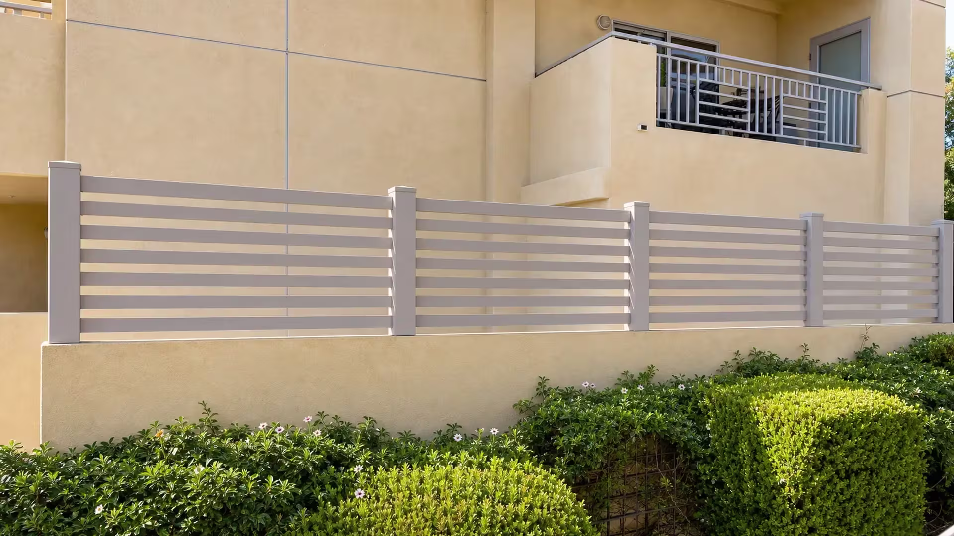Clay vinyl picket fence below a balcony rail, framed by clipped shrubs and flowering beds