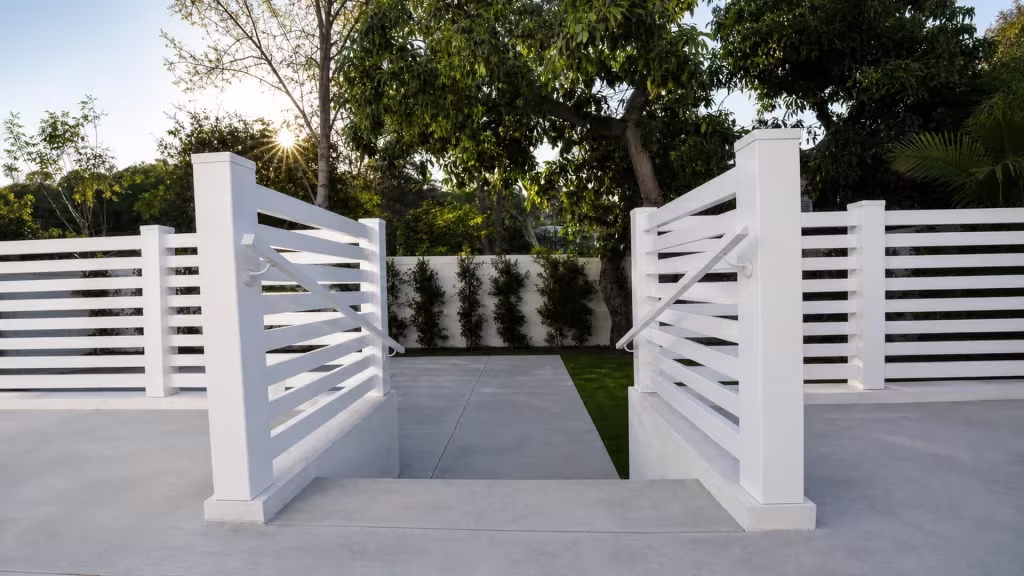 A White vinyl picket fence frames a double swing gate opening onto a concrete walkway