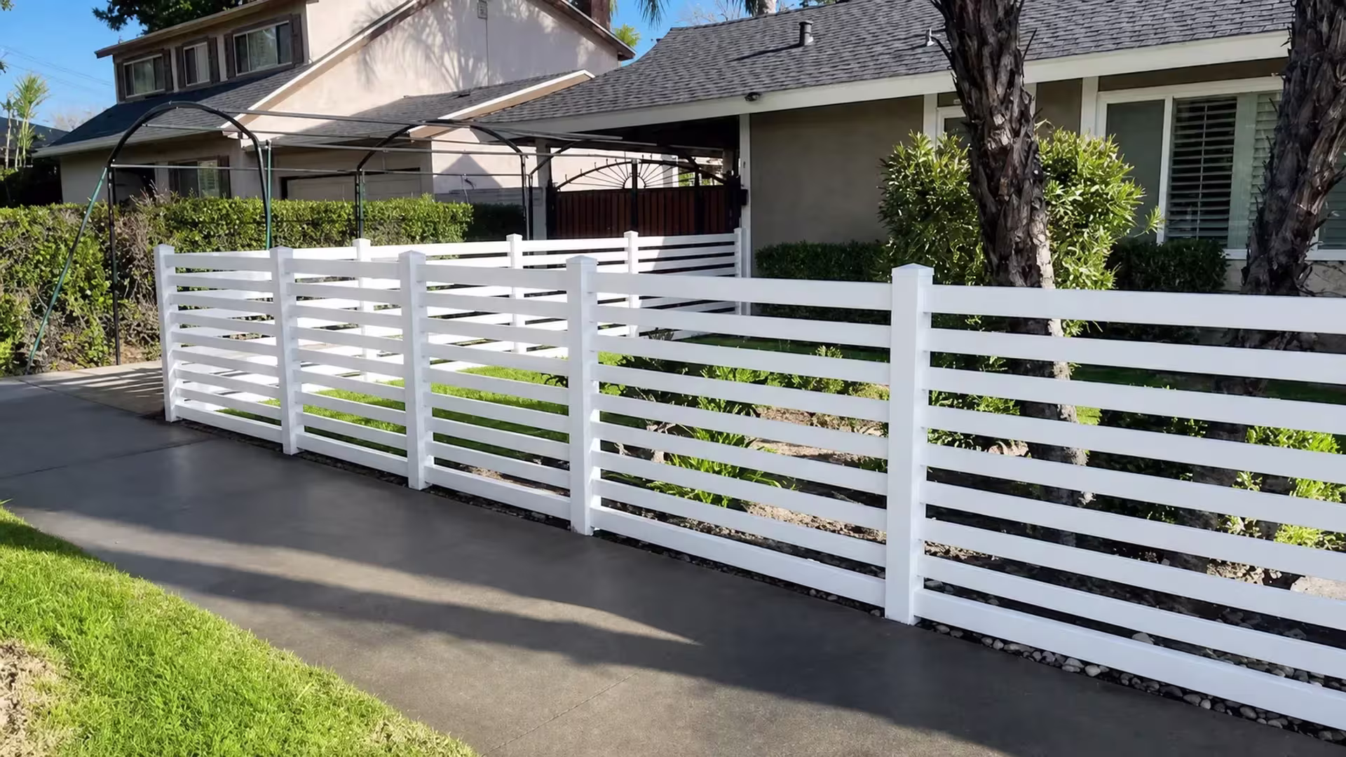 Straight White vinyl horizontal picket fence marks a front yard boundary past palm trunks and a gravel strip