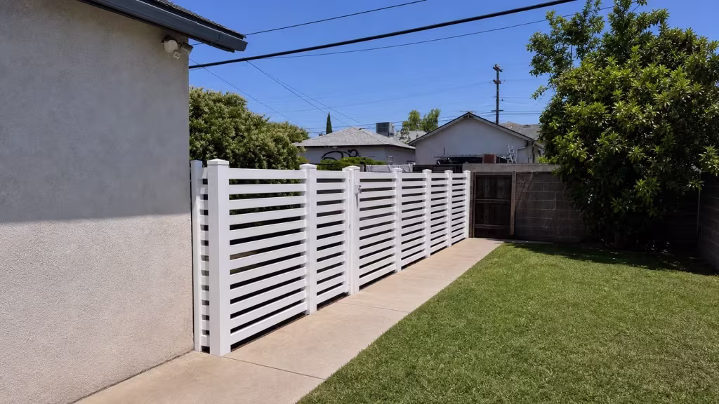 White vinyl closed-top picket fence recedes beside a house wall, with existing gate latch hardware left mid run