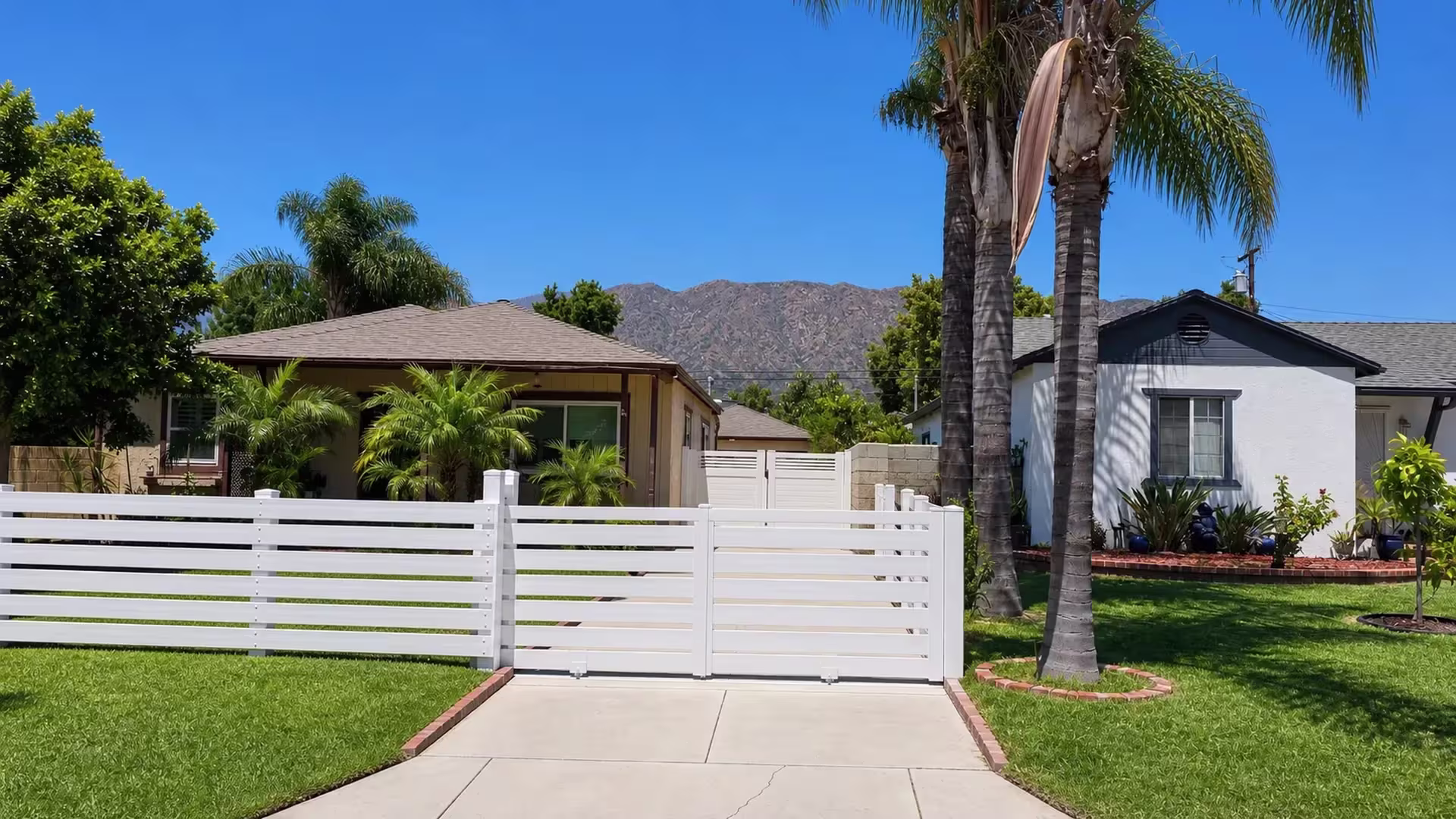 A two-leaf driveway gate joins this White vinyl picket fence across the front of the property beneath a mountain backdrop