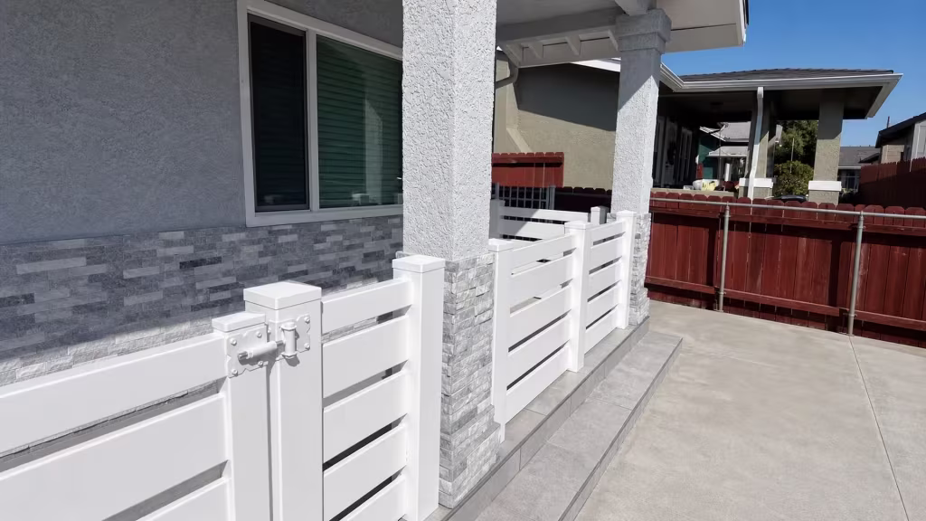 A raised tiled step carries this White vinyl picket fence and gate past the same stacked-stone columns