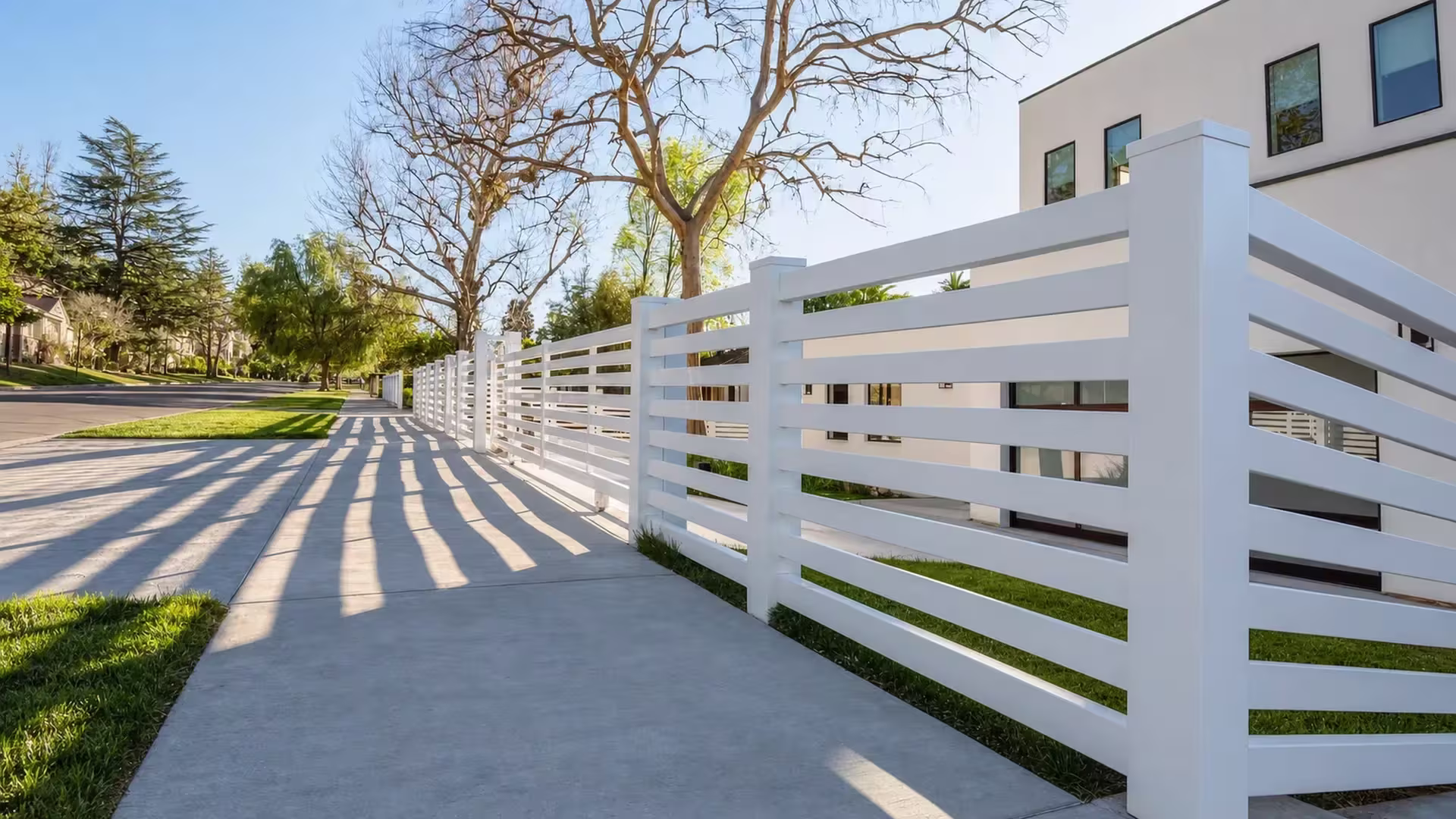 Long White vinyl picket fence casting late shadows across a concrete sidewalk near a modern house