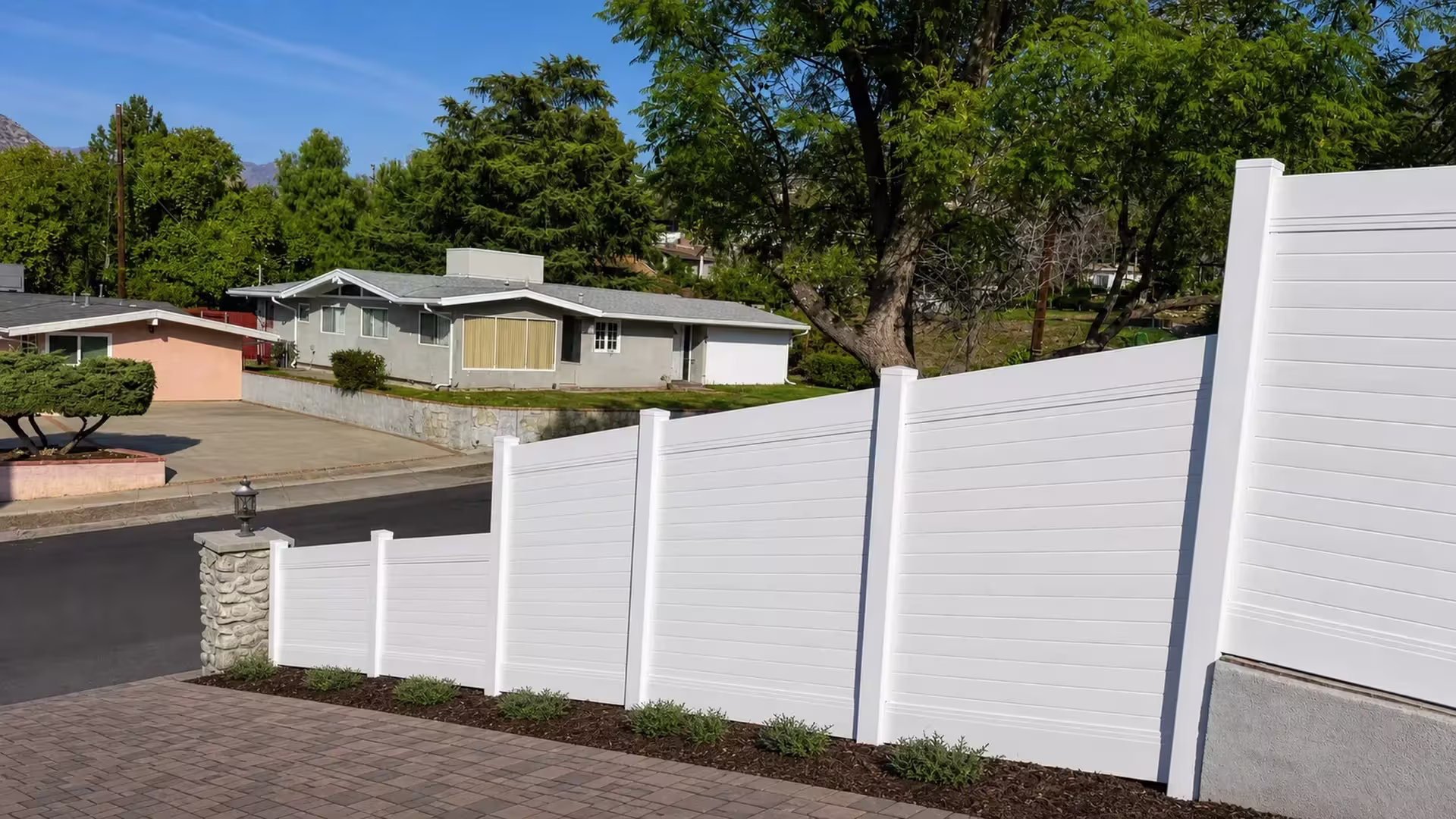 A stone column marks the start of a White vinyl horizontal privacy fence along a sloped boundary