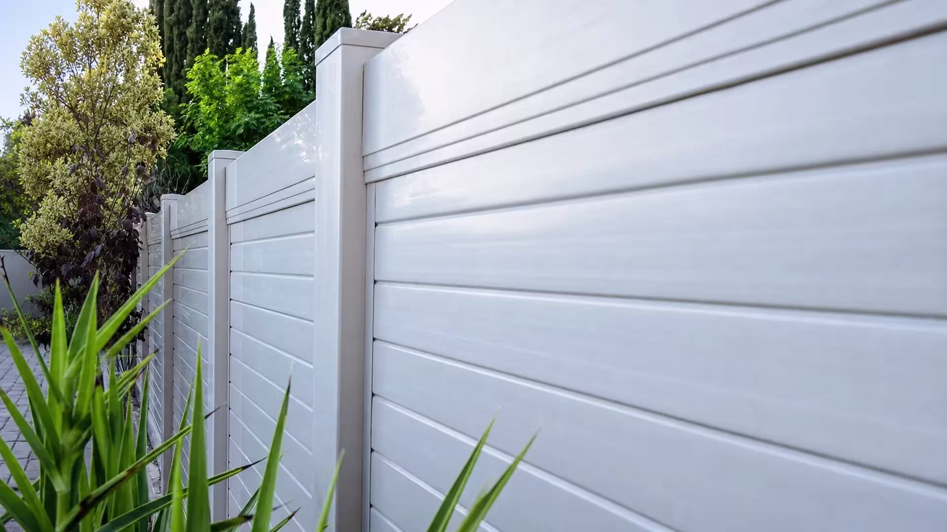 Established trees and shrubs fill the yard behind a White vinyl horizontal privacy fence lined with sword-like foliage