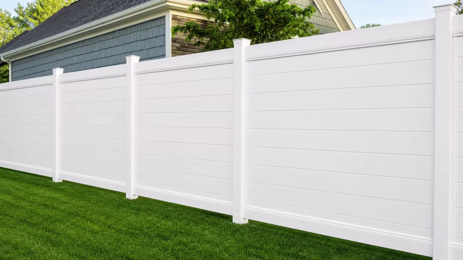 A residential lawn is crossed diagonally by a White vinyl horizontal privacy fence past a stone veneer wall