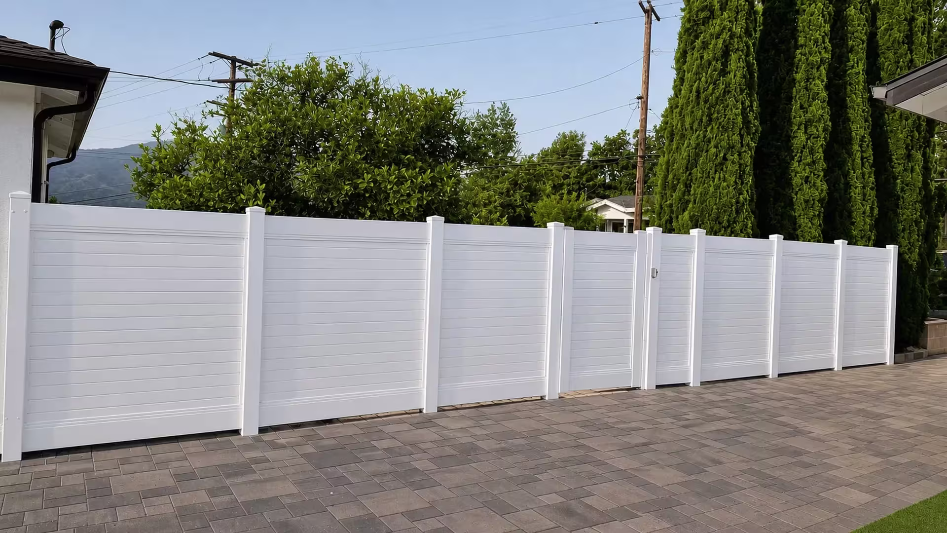 A paved residential driveway is spanned by a White vinyl horizontal privacy fence near distant mountains