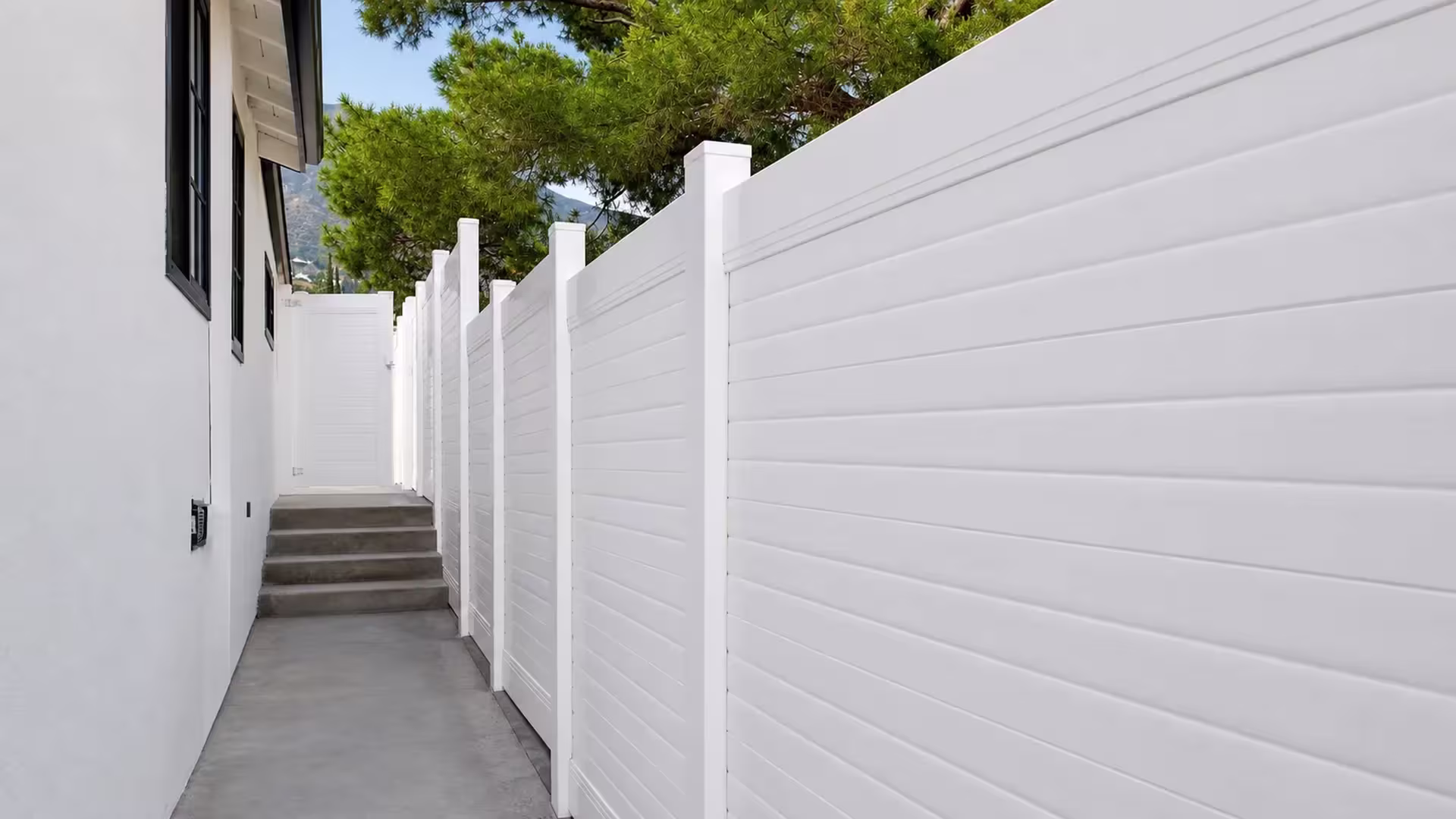 A rising side yard grade is followed by a White vinyl horizontal privacy fence toward a distant mountain glimpse