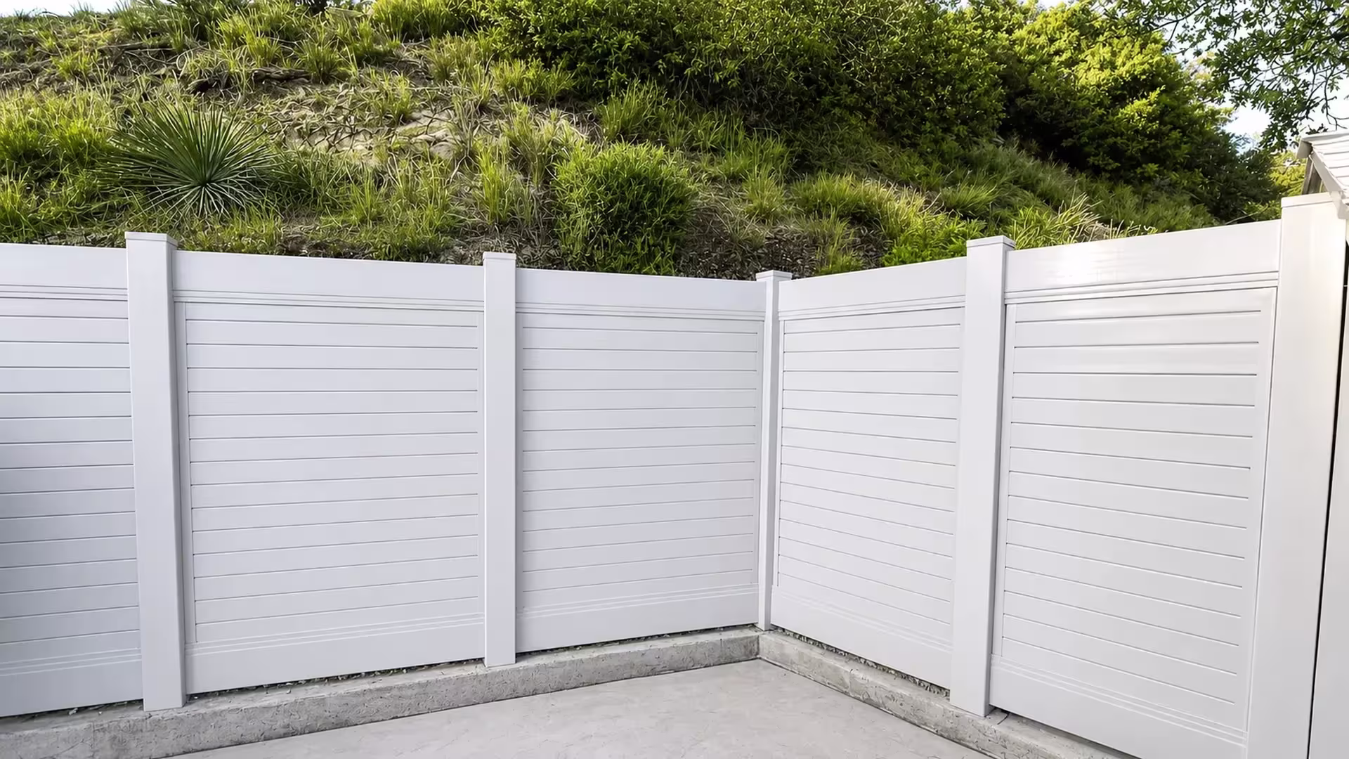 A concrete patio corner is wrapped by a White vinyl horizontal privacy fence on hillside terrain