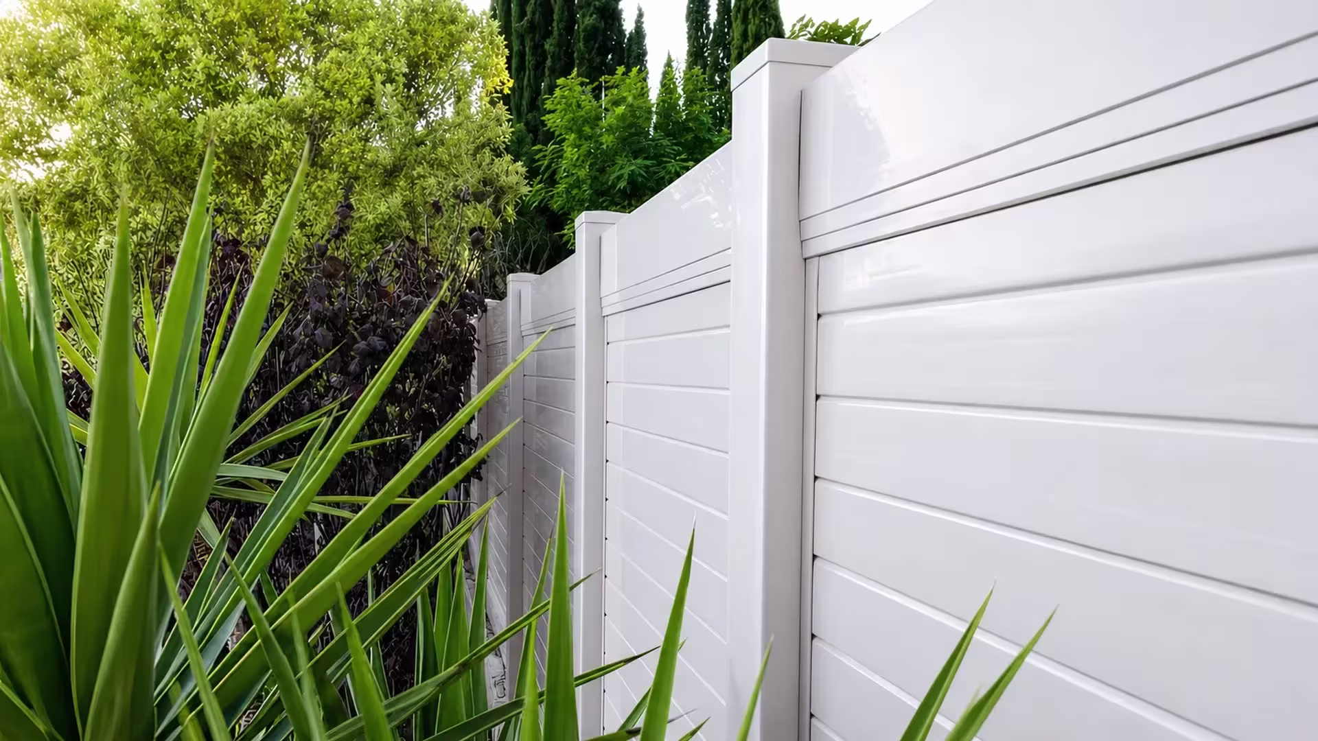 Burgundy hedge and cypress trees surround a White vinyl horizontal privacy fence through dense landscaping