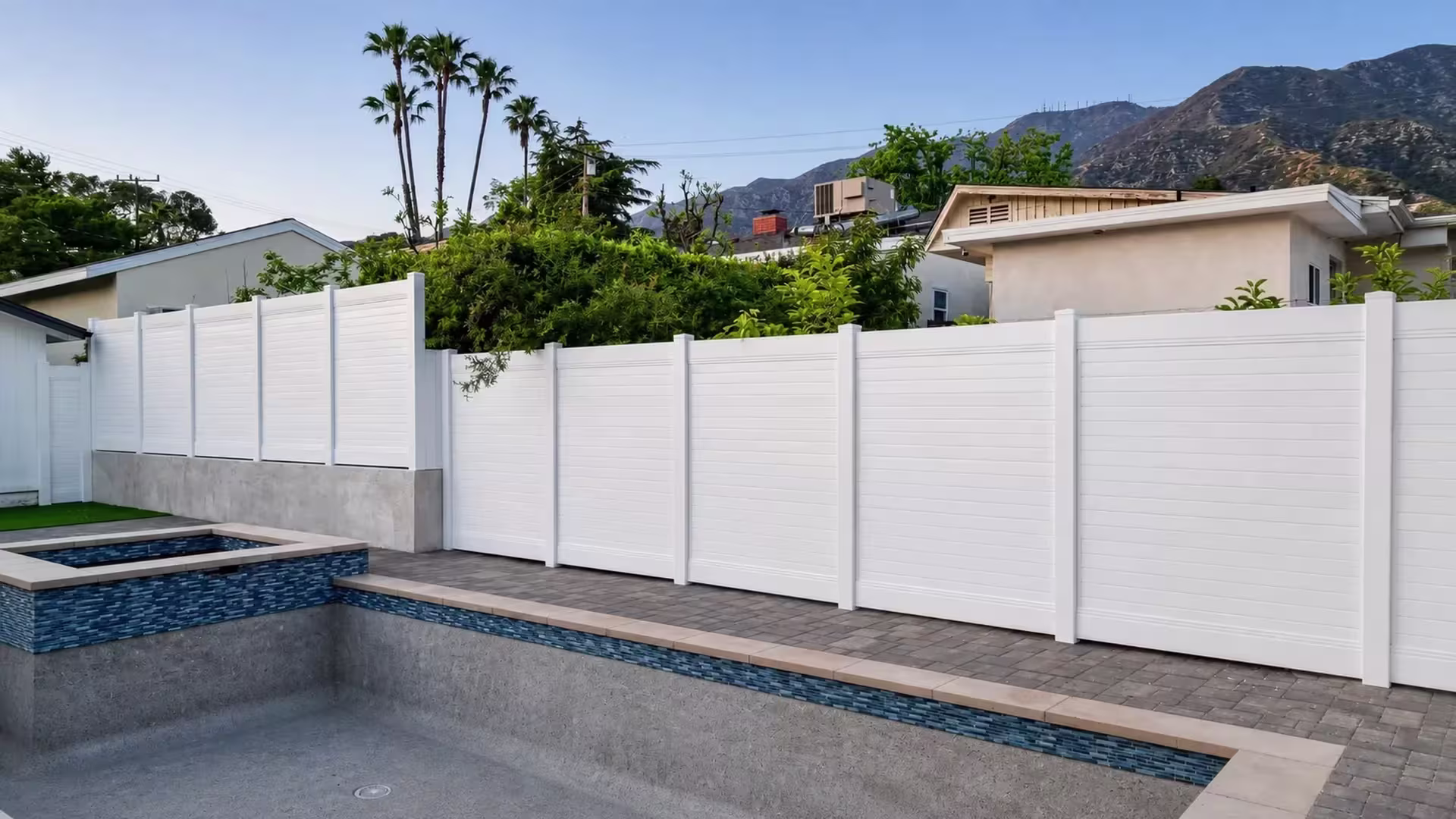 A backyard pool deck is enclosed by a White vinyl horizontal privacy fence along a stepped wall