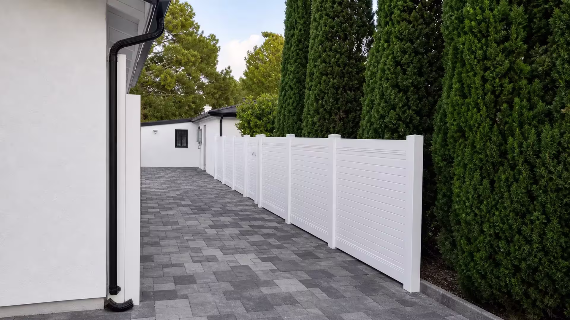 A tall evergreen hedge runs beside a White vinyl horizontal privacy fence along the paver walkway