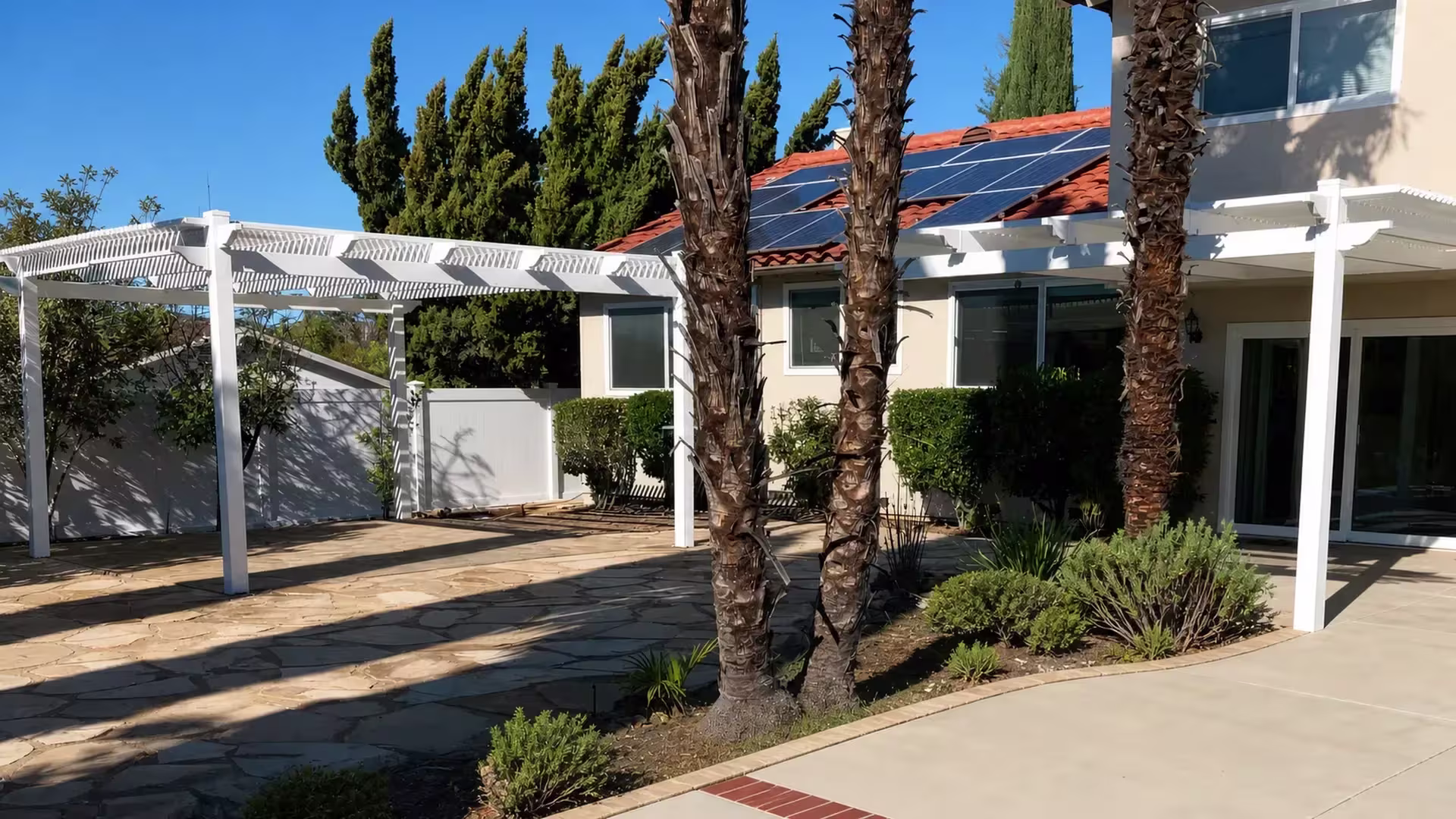 A solar-paneled tile roof on the stucco house rises behind an open lattice White vinyl pergola