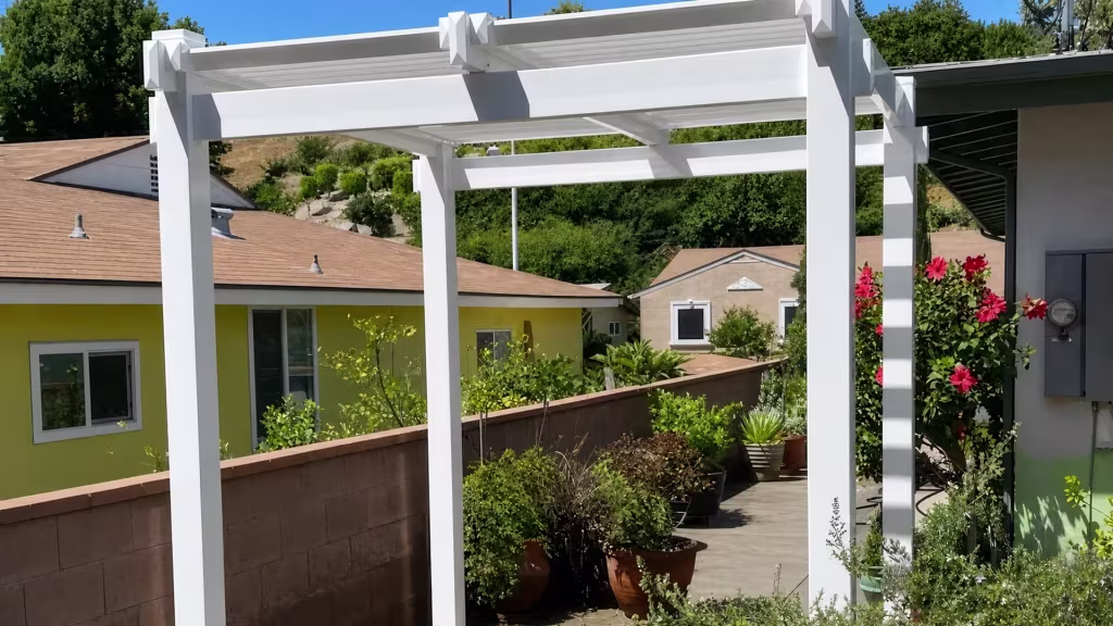 A hillside lot behind a masonry wall carries a White vinyl pergola on four square posts with rafter ends