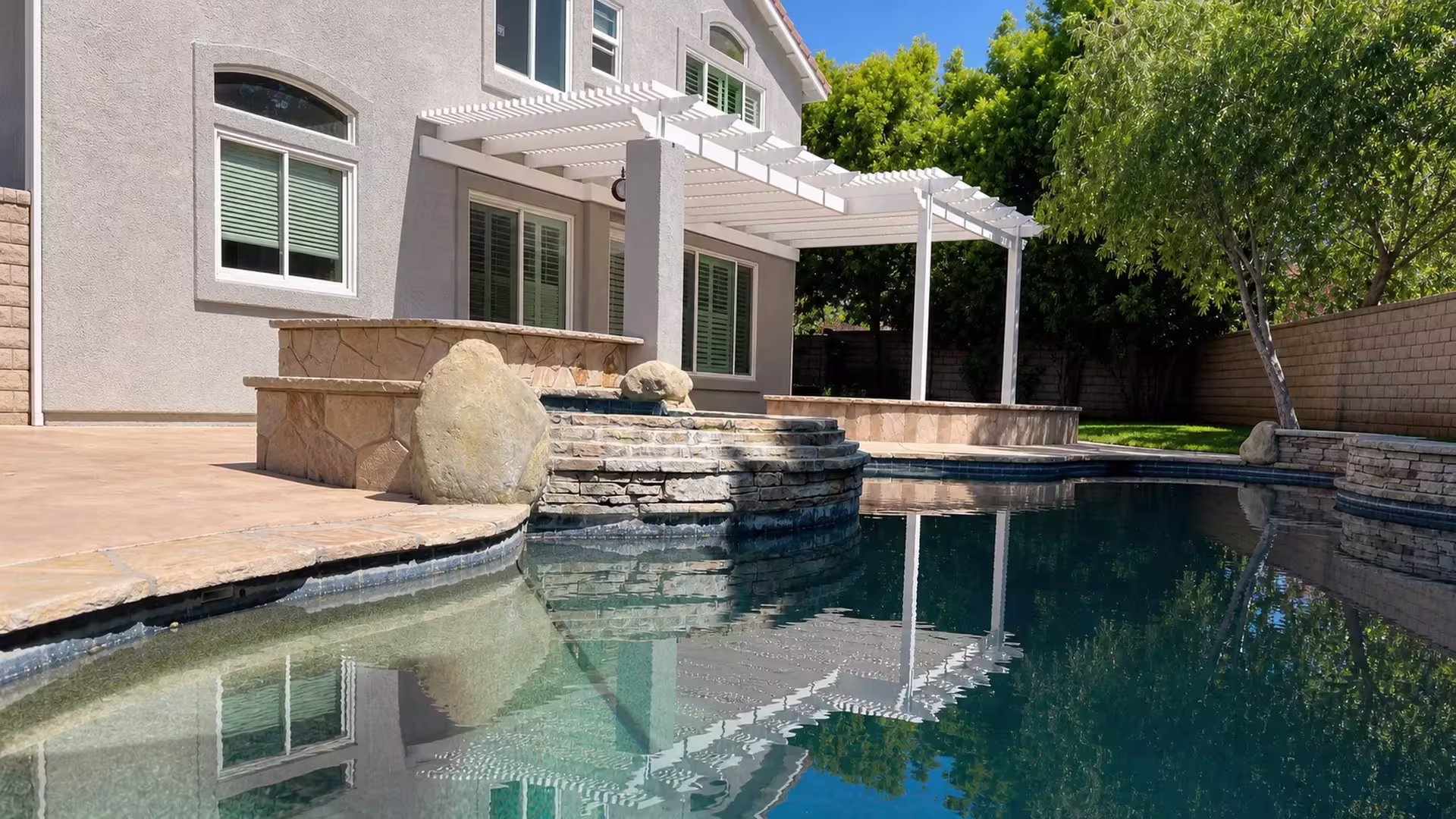A raised stone spa with a spillway sits just below a White vinyl picket patio cover