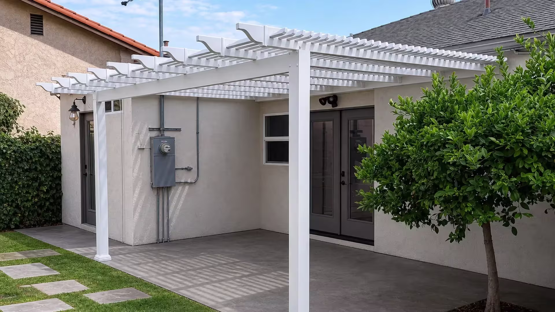 White vinyl open-lattice picket patio cover shading a paved patio beside a mature citrus tree