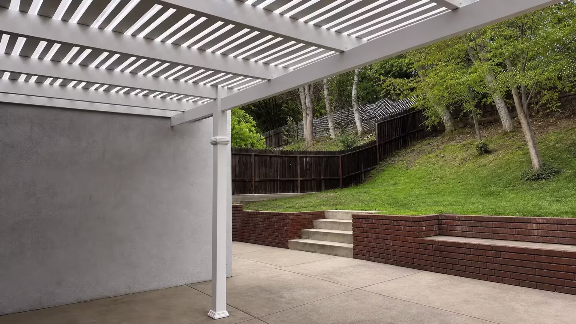 Sloped yard and brick retaining walls meet a White vinyl picket patio cover with an open lattice roof
