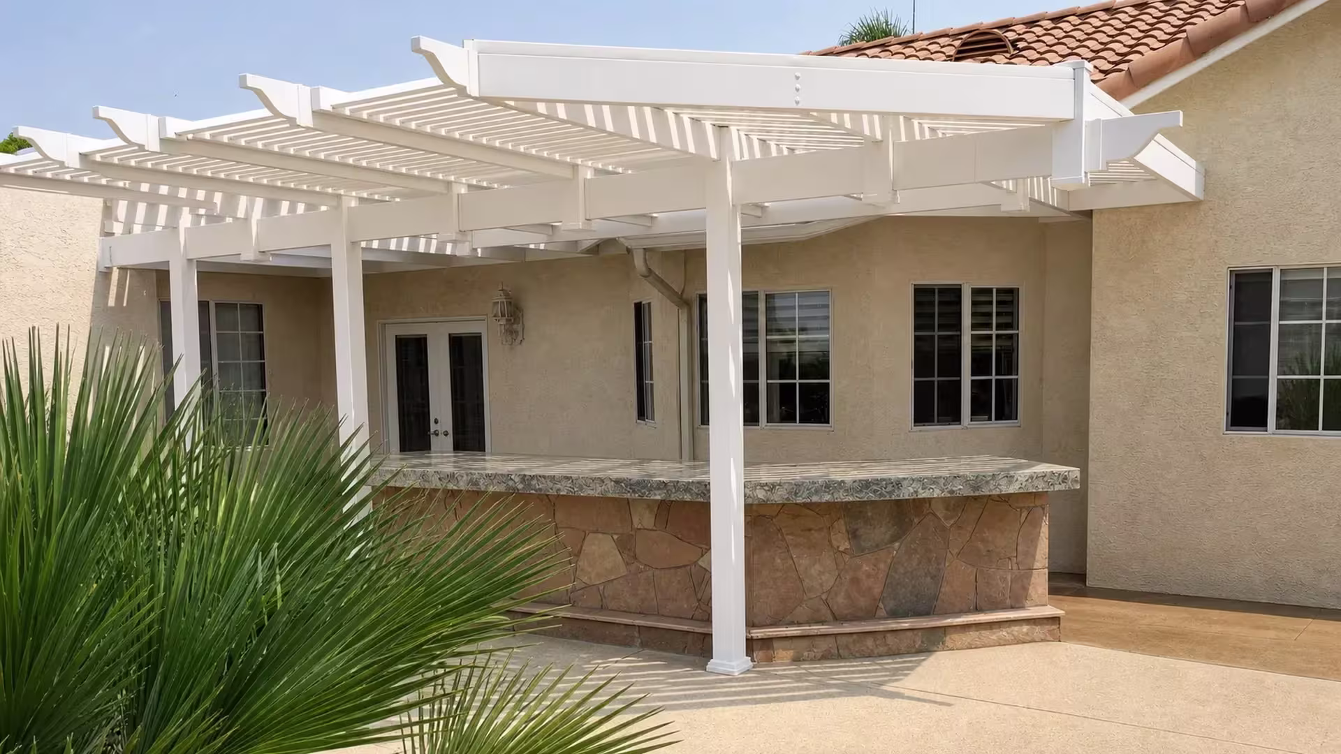 An outdoor bar counter stands beneath a White vinyl picket patio cover topped by a clay tile roofline