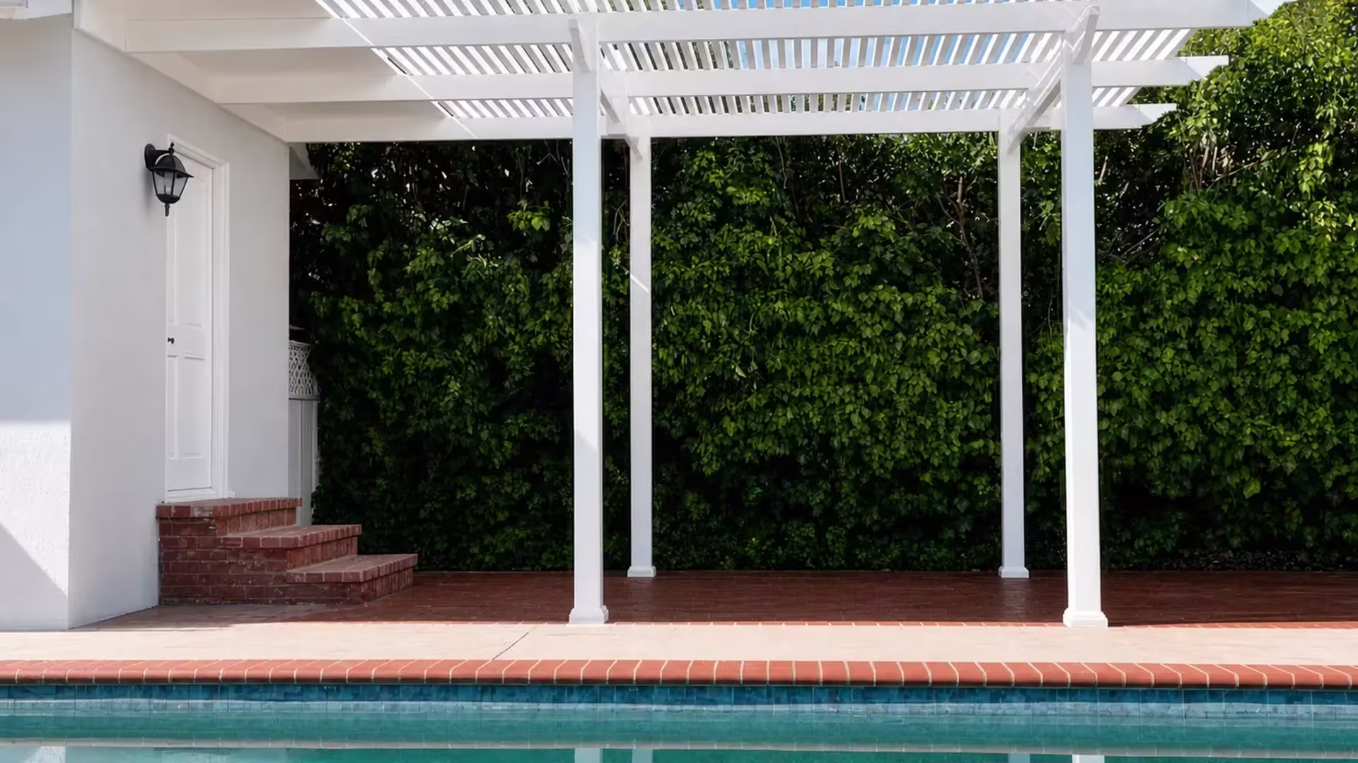 A poolside wall with a black lantern and brick steps sits next to this White vinyl picket patio cover