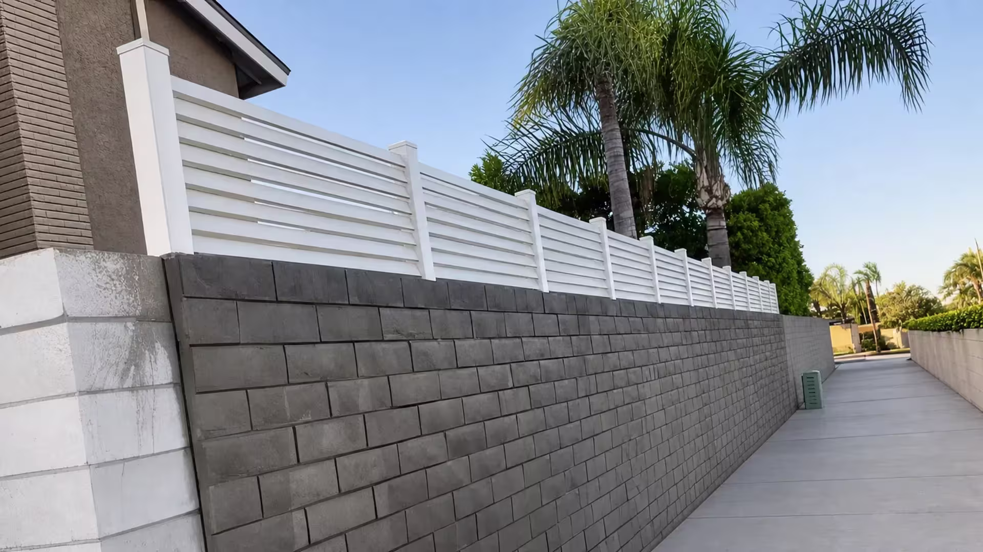 White vinyl picket wall topper sits atop a gray block wall below a neighboring house roofline and hedge