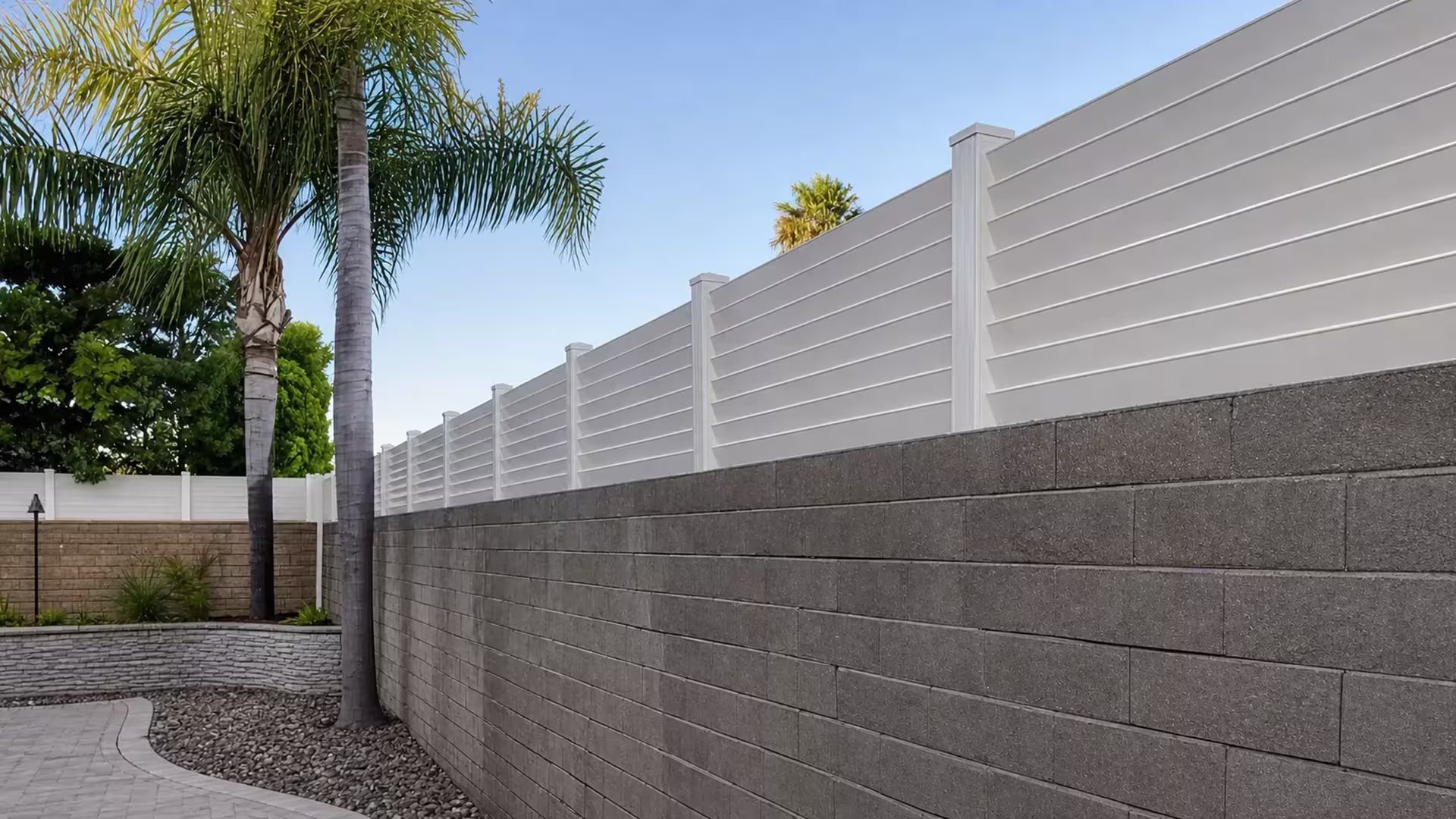 Seven-panel run of White vinyl picket topper spans a gray masonry wall past palms, trees, and shrubs