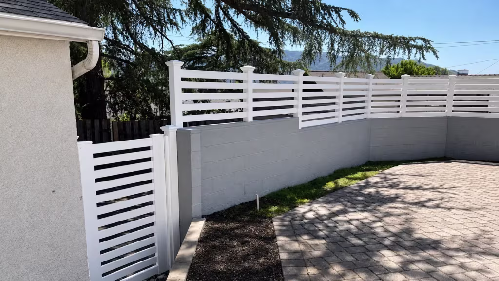 Painted masonry wall beside a paved courtyard carries a White vinyl picket topper under distant hills