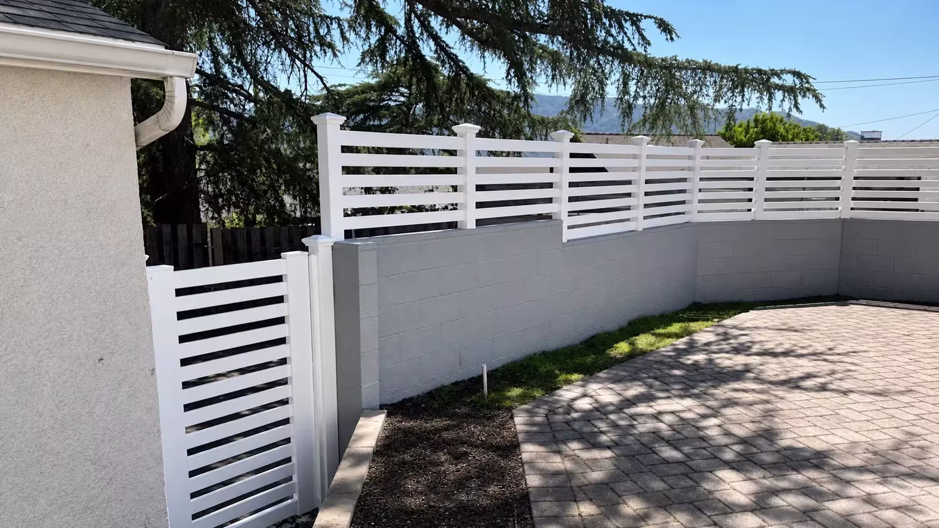Painted masonry wall beside a paved courtyard carries a White vinyl picket topper under distant hills