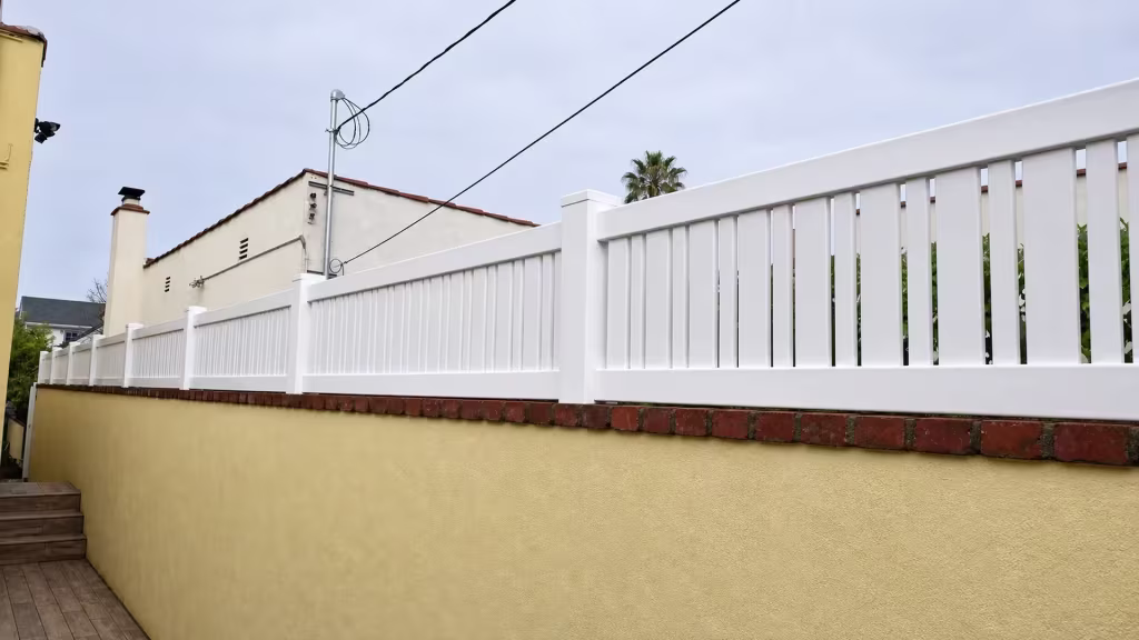 Pale yellow stucco wall carries a White vinyl picket topper along a narrow walkway and steps