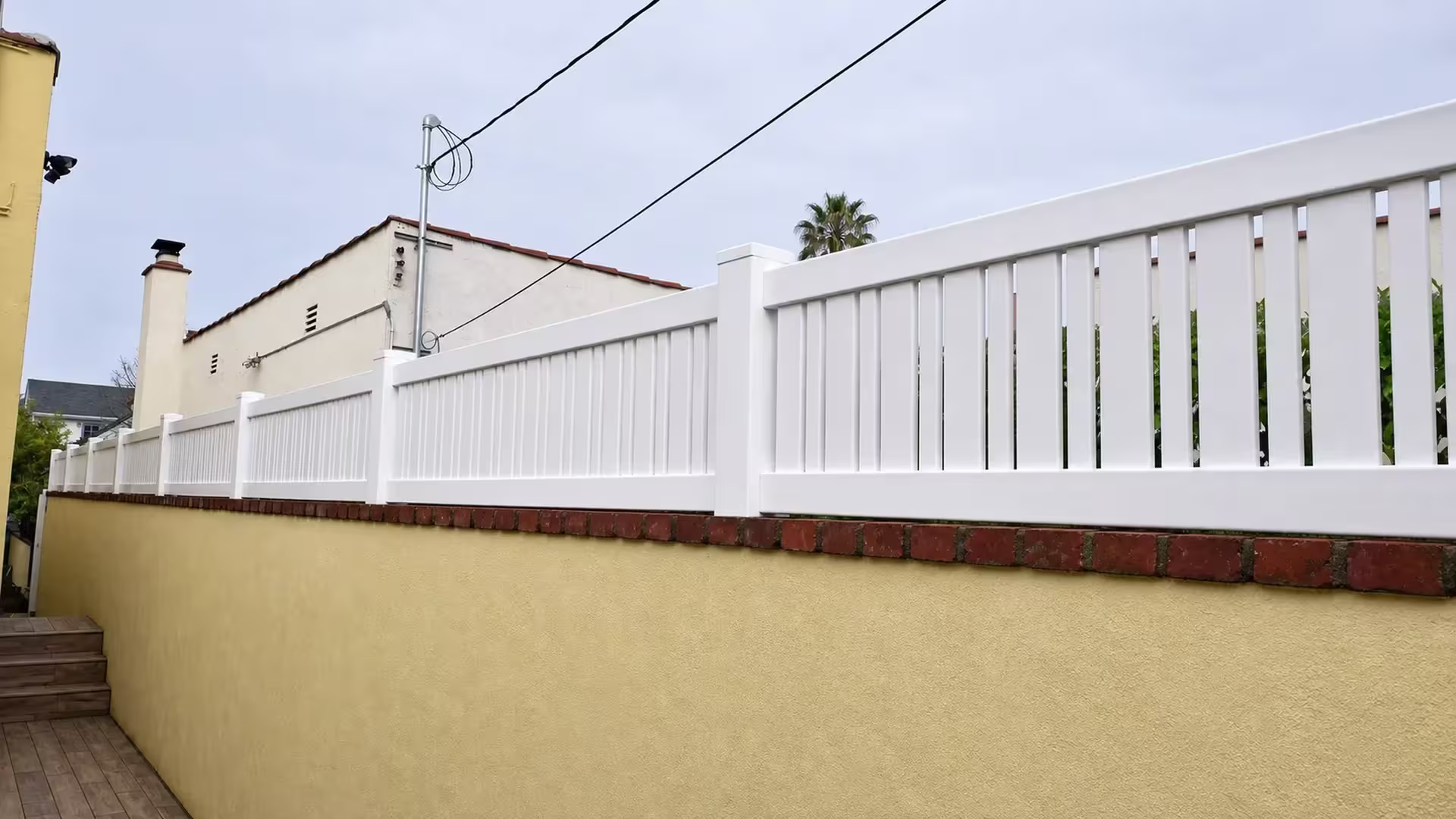 Pale yellow stucco wall carries a White vinyl picket topper along a narrow walkway and steps