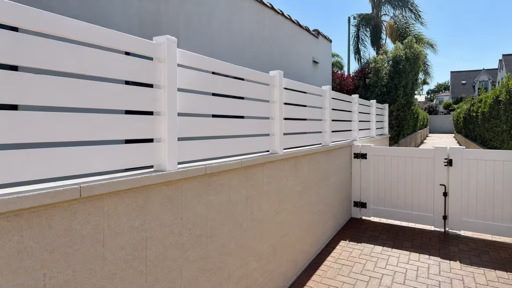 White vinyl picket wall topper runs along a beige block wall toward a paved driveway