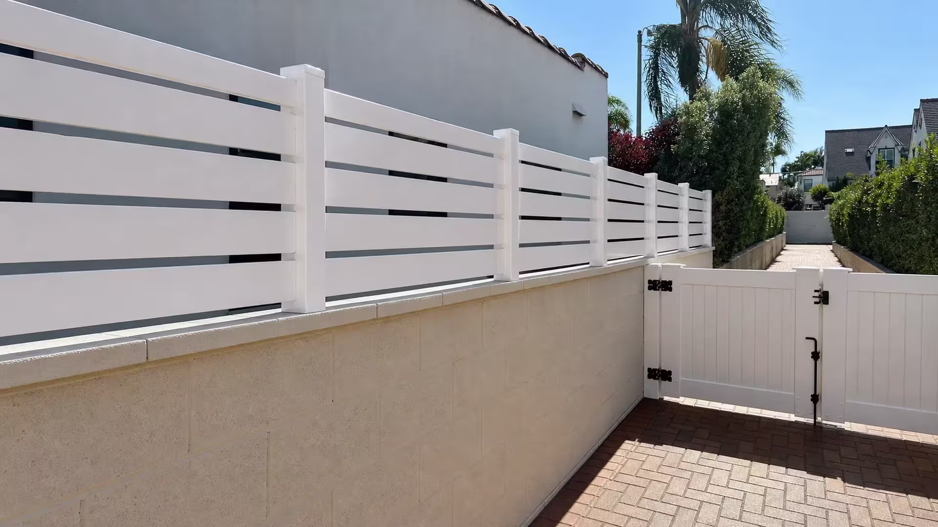 White vinyl picket wall topper runs along a beige block wall toward a paved driveway