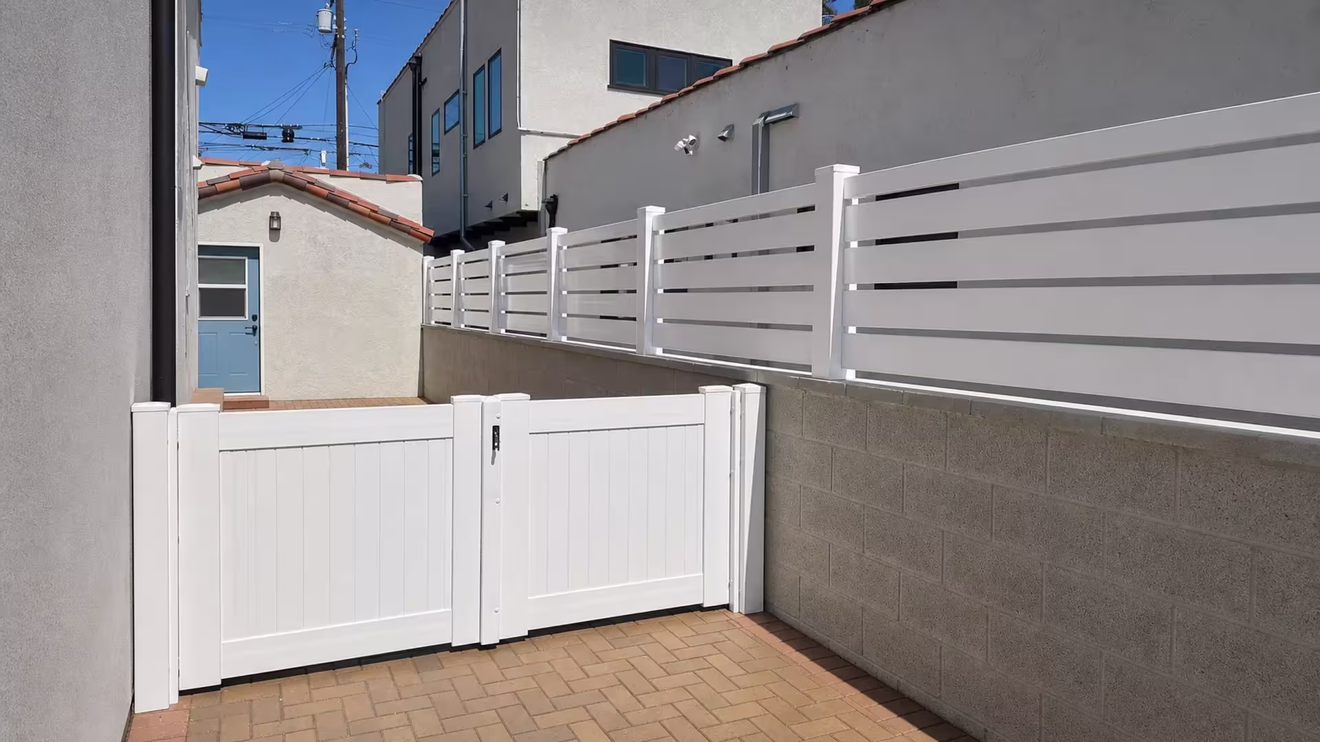 Matching gate on a brick paver walkway stands beside a White vinyl picket wall topper near stucco buildings