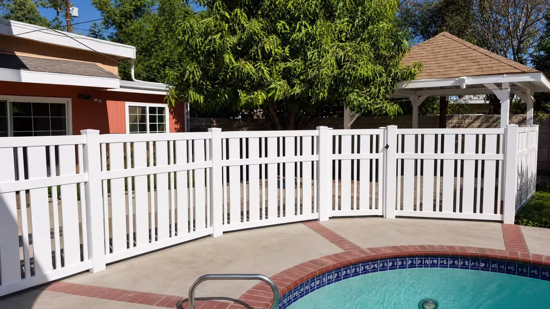 White vinyl close-top picket pool fence with a single hinged gate set between fixed panels near a garden gazebo