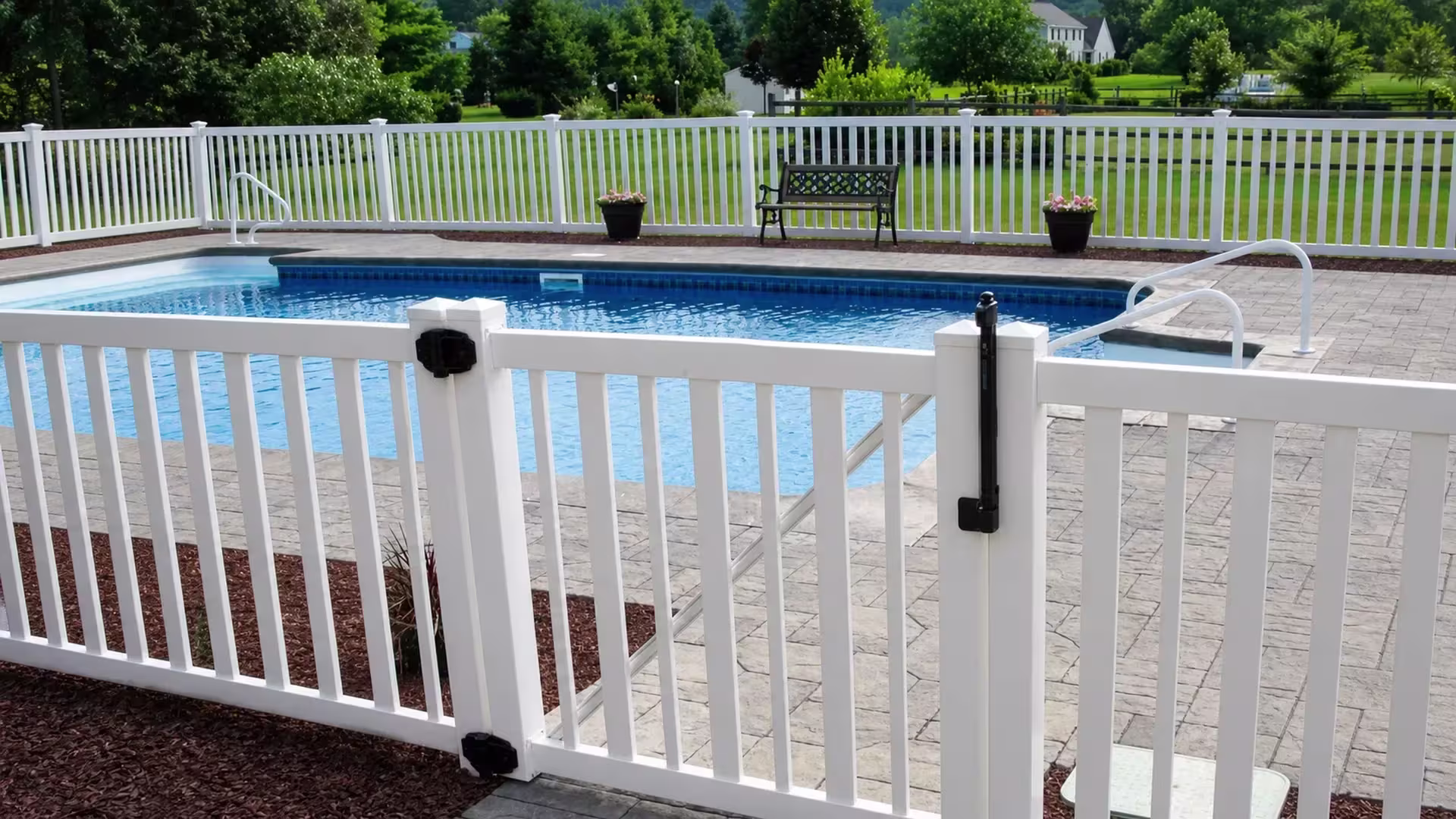 An inward-facing gate leaf on a White vinyl pool fence swings from a stamped-concrete deck near a bench and planters