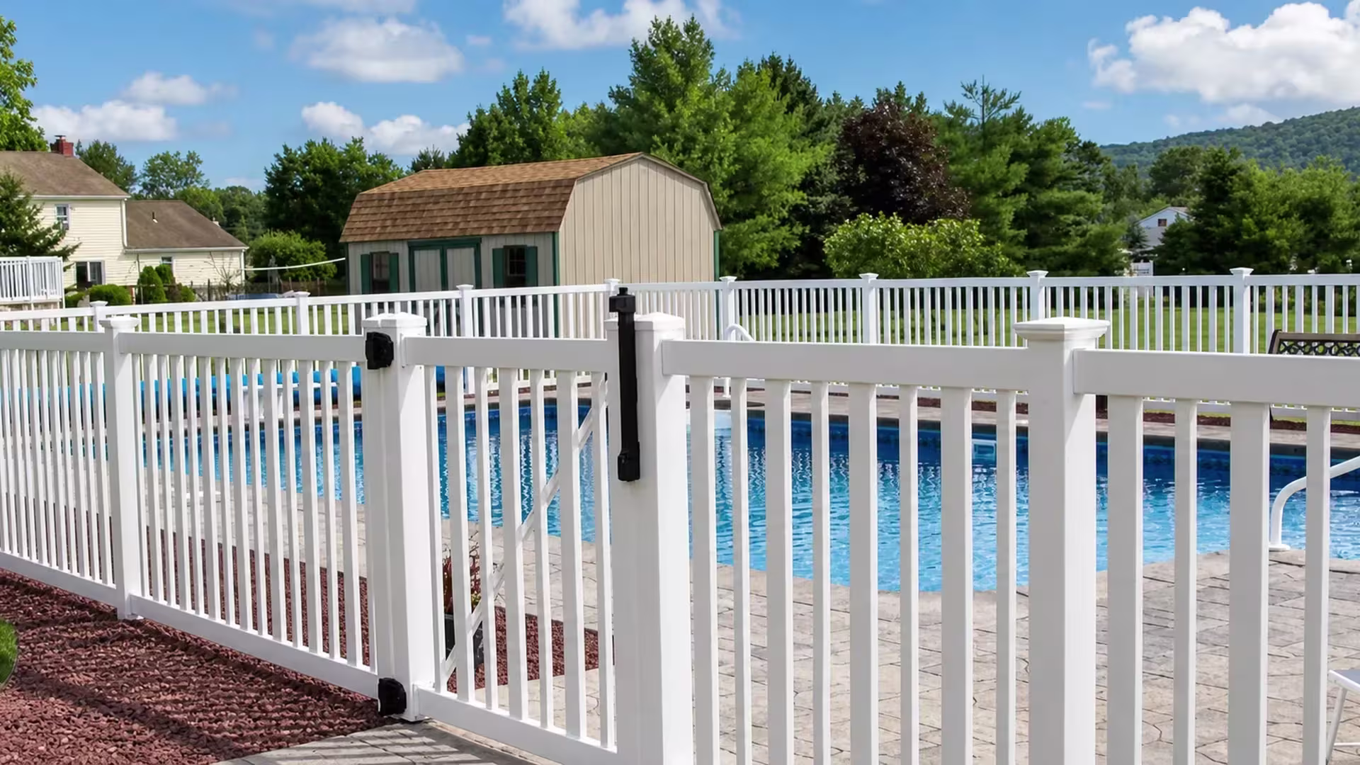 Beige vinyl pool fence with a foreground picket gate and a distant wooded ridge rising behind the property