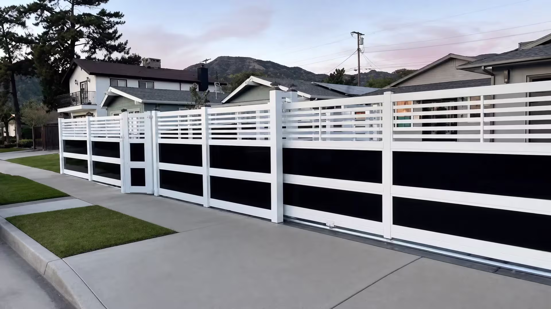A narrow pedestrian gate opens this White vinyl privacy fence with acrylic beside a strip of front lawn