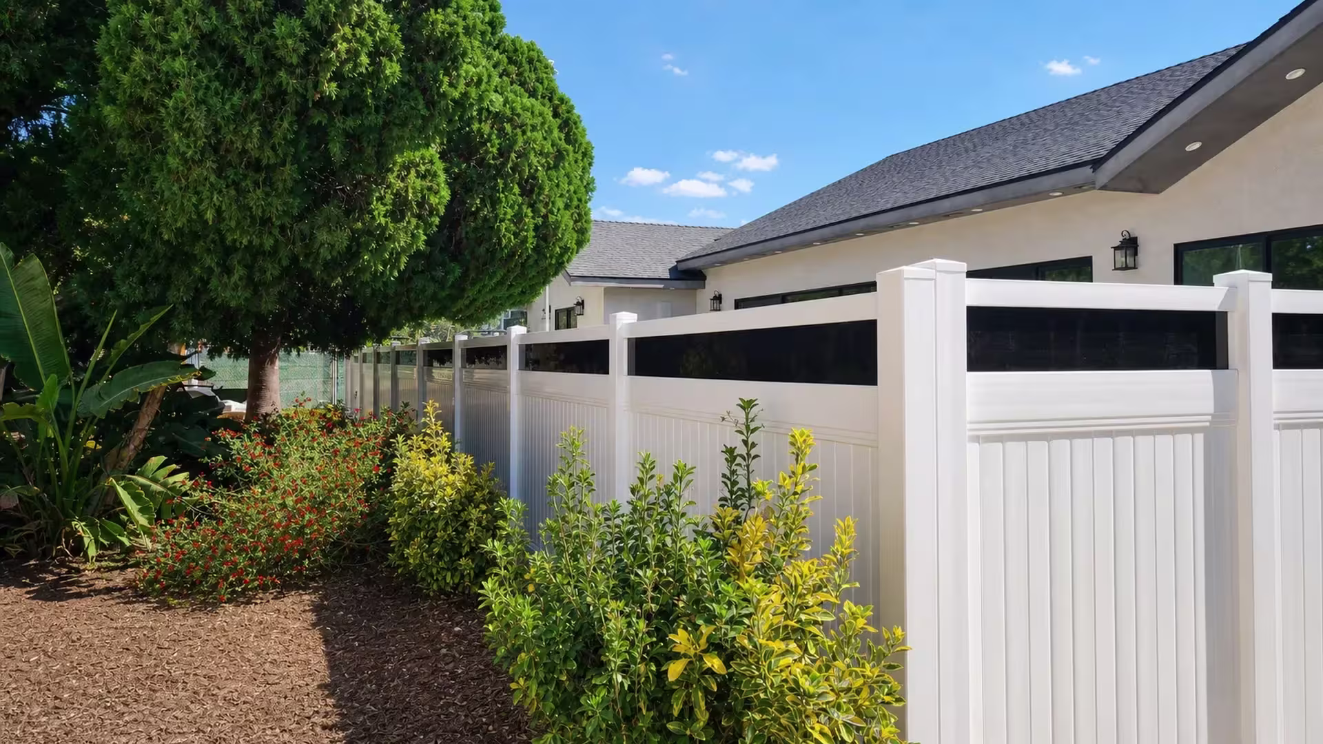 A cream stucco house borders this White vinyl privacy fence with acrylic angling toward five pyramidal-capped posts