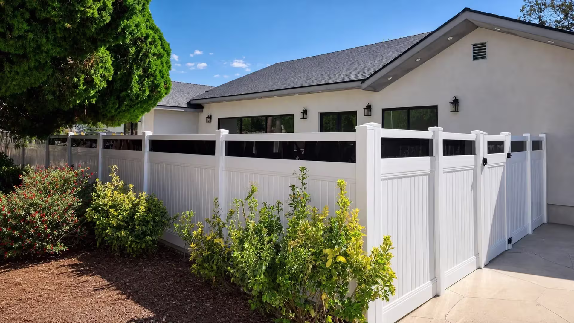 A mature evergreen tree shades this White vinyl privacy fence and gate with acrylic guarding a light stucco house
