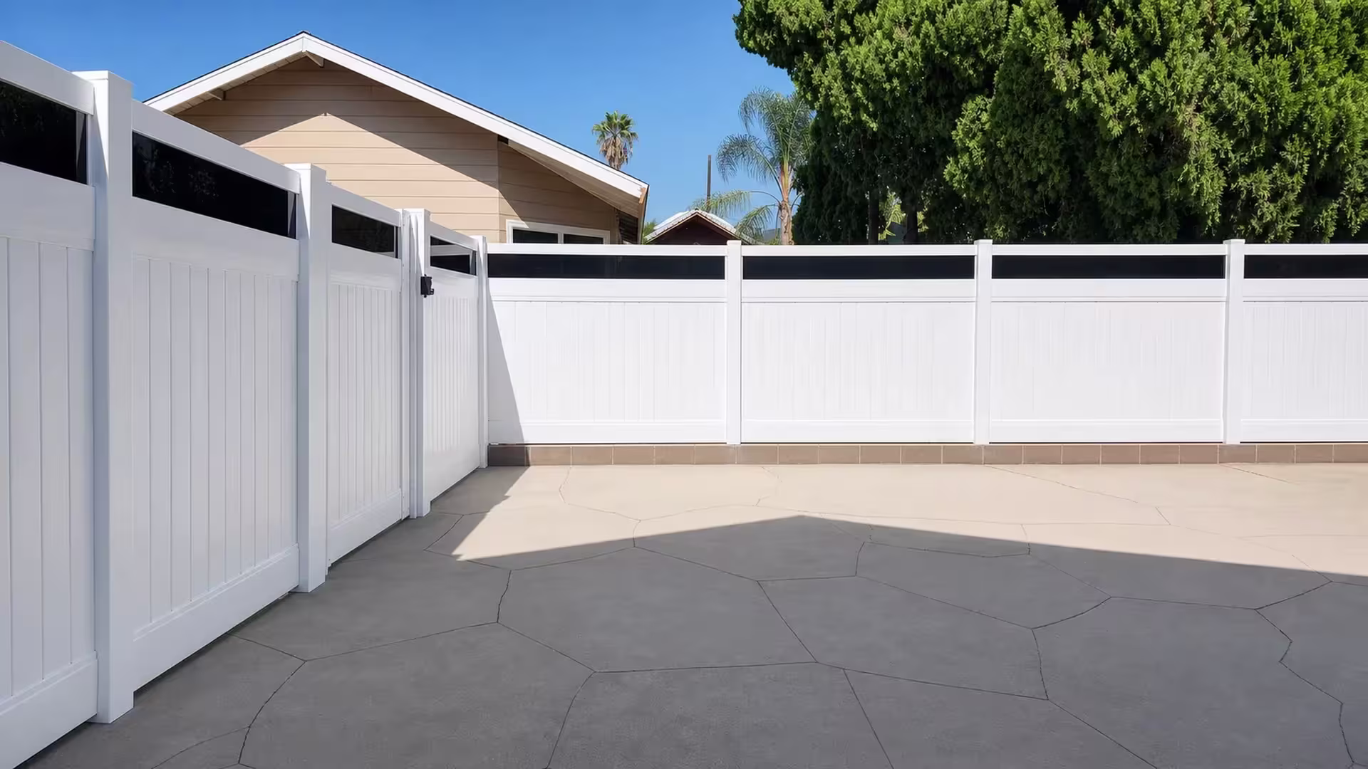 A tan gabled house stands beside this White vinyl privacy fence with acrylic bending past a cracked concrete patio