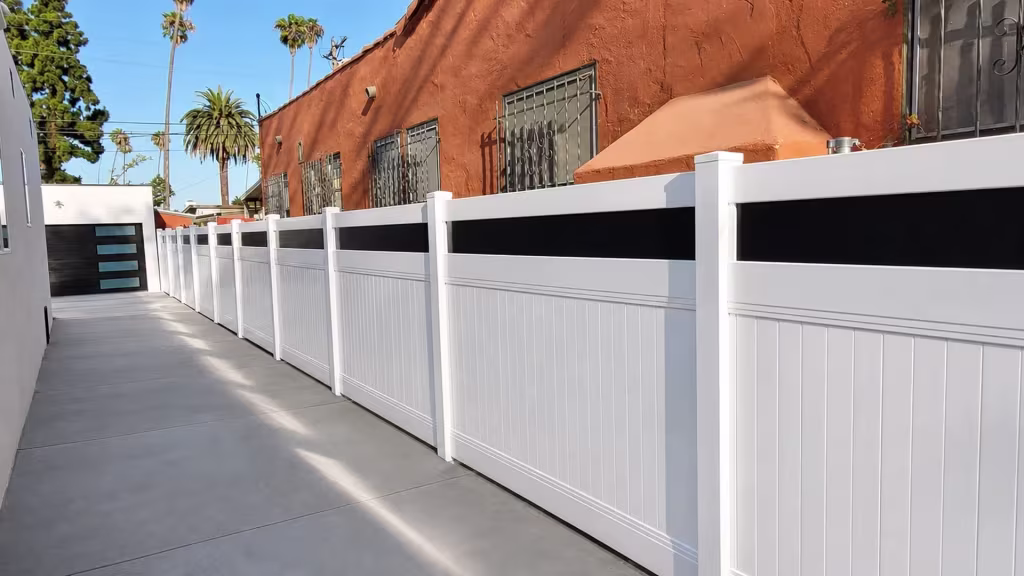 A terracotta stucco wall with barred windows lines this White vinyl privacy fence with acrylic leading to a detached garage