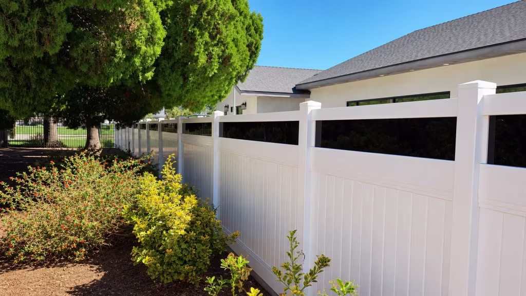 A dark gray shingle roof shows behind this White vinyl privacy fence with acrylic sitting beneath mature evergreen trees